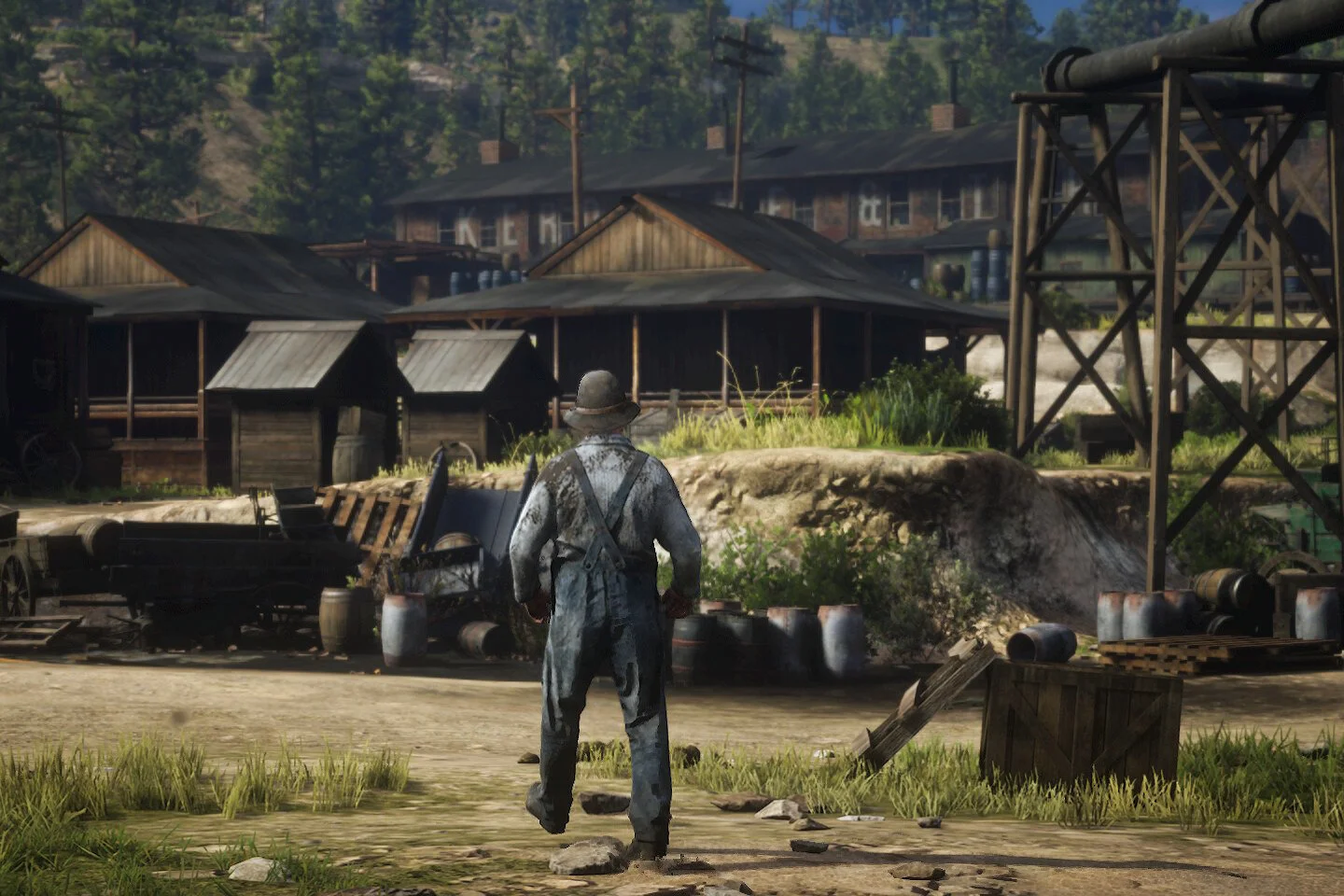 A person wearing worn, dirty clothes and a hat walks through an abandoned, slightly overgrown industrial area with wooden buildings, barrels, and scattered debris, with hills and trees in the background.