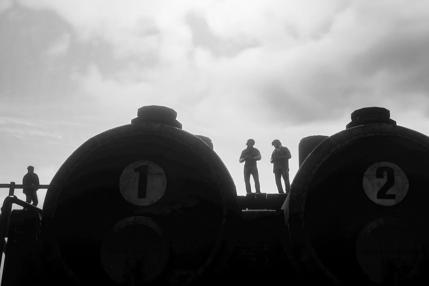 Silhouettes of five people on a rooftop or platform with two large industrial tanks marked '1' and '2' against a cloudy sky.