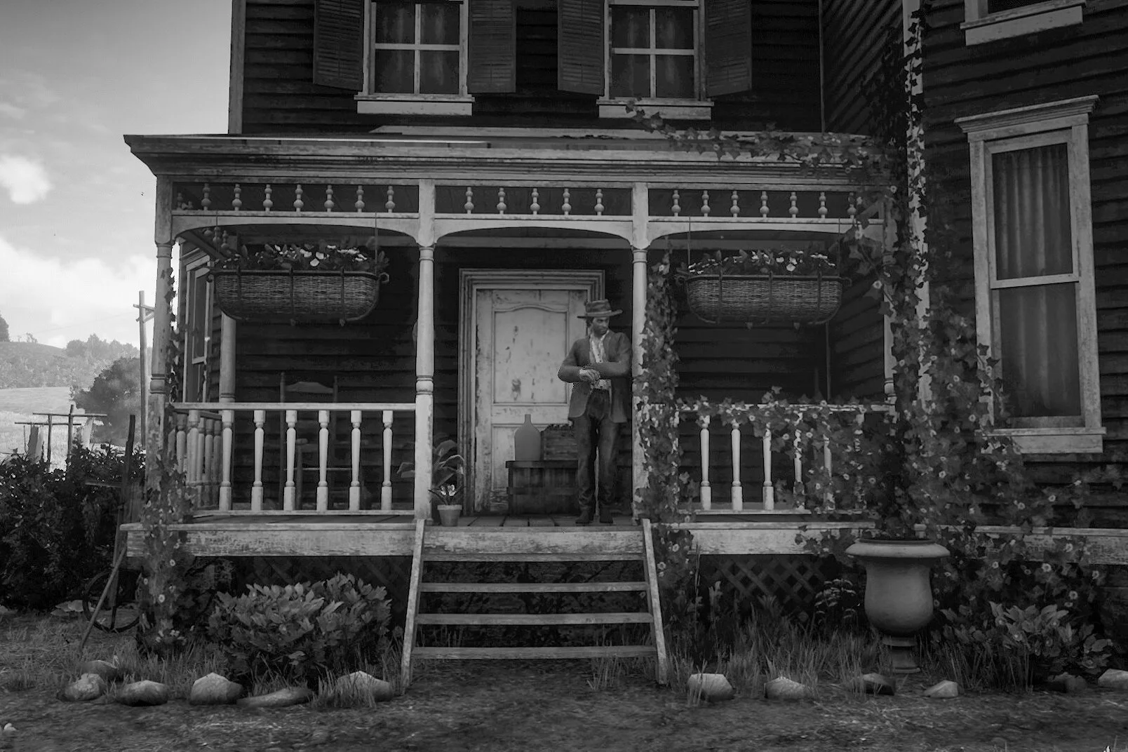 A black-and-white photo of a man standing on the porch of a wooden house, dressed in period clothing with a hat, next to a large ceramic vase. The house has a staircase leading up to the porch, with potted plants and hanging baskets, and ivy climbing