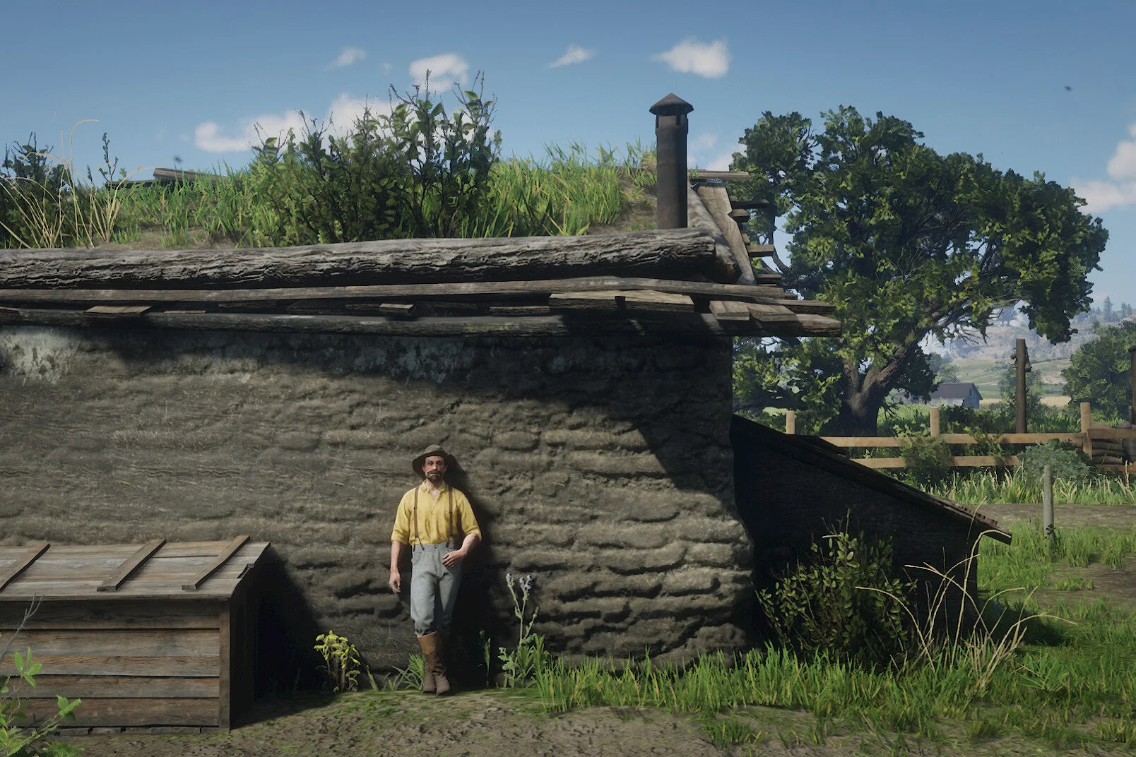 A man standing against a rustic stone wall with a grassy rooftop and a large tree in the background, in a rural setting.