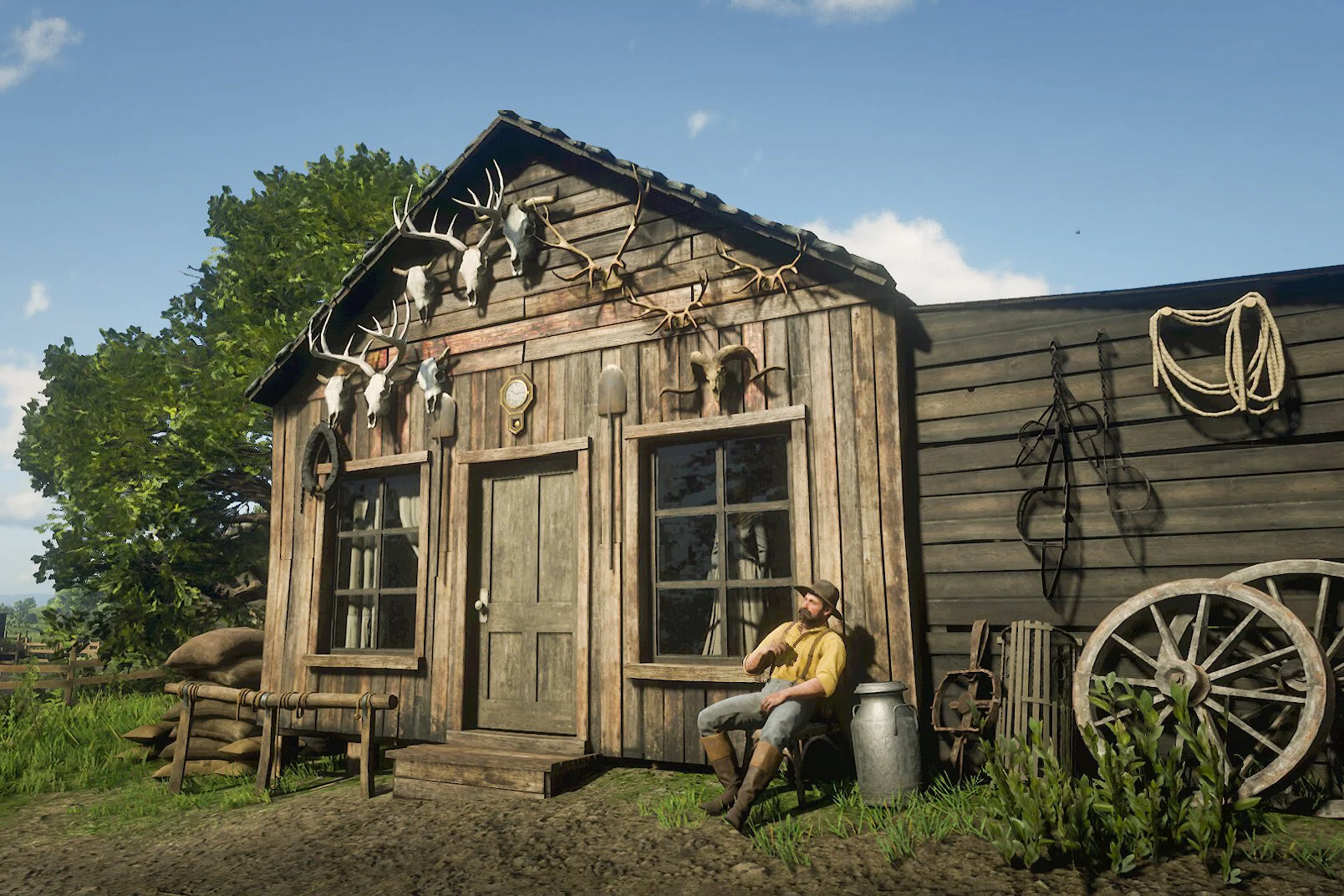 A rustic wooden farm shed with antlers and skulls mounted on the wall, a man in vintage clothing sitting outside, surrounded by farming tools, a large water pail, and a wagon wheel, with a tree and a blue sky in the background.