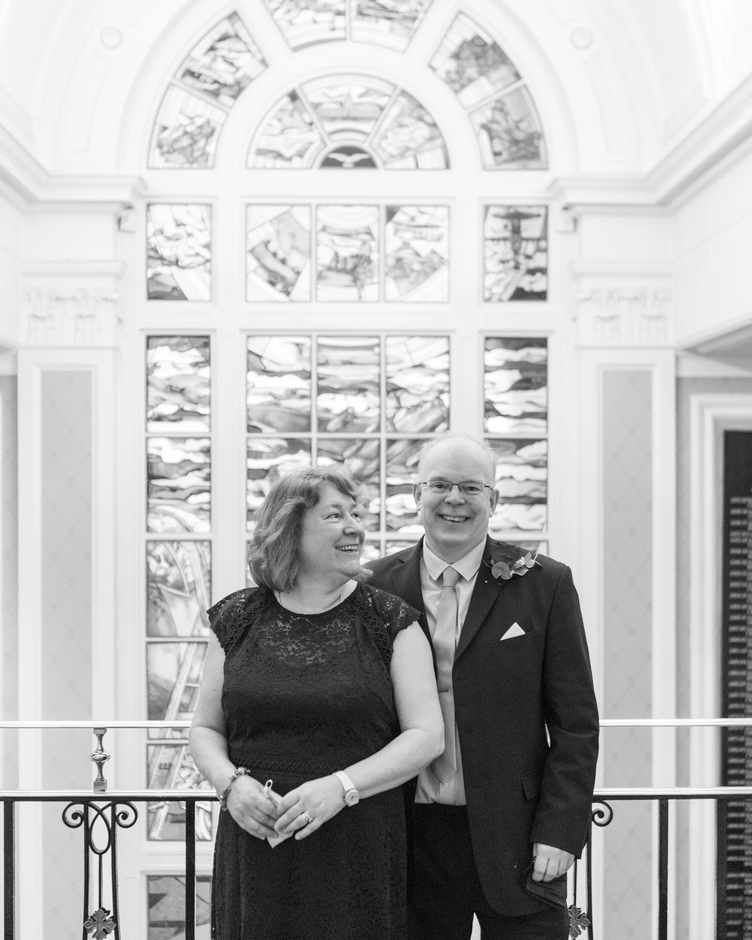 A smiling woman and man dressed in formal attire standing together in front of a stained-glass window.