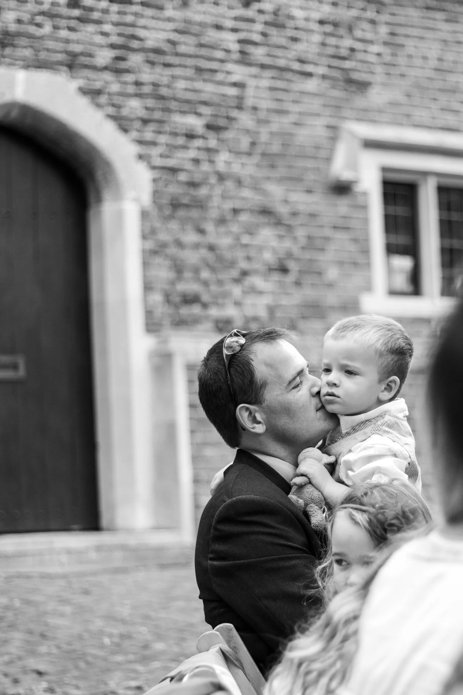 A man is kissing a young boy on the cheek at an outdoor gathering, with other children nearby. The scene is in front of a brick building.