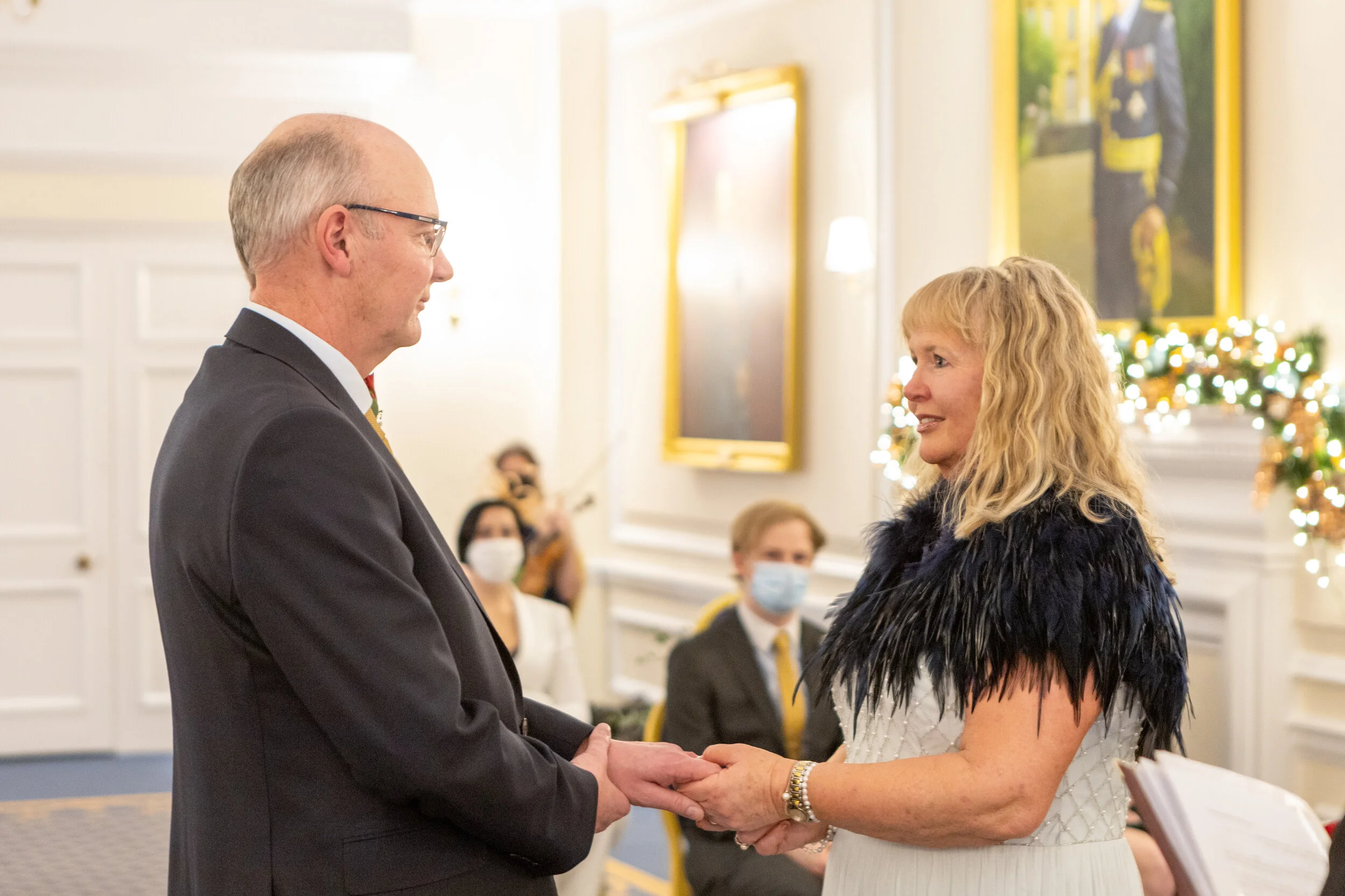 A man and woman holding hands and looking at each other during a wedding ceremony inside a decorated room with people watching in the background.