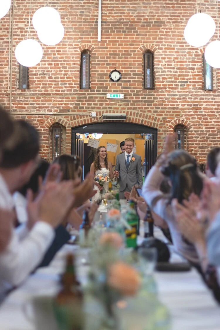 A wedding reception with a bride and groom walking into the room, smiling and holding hands. The room has a brick wall and hanging lights. Guests are sitting at a long table, clapping and cheering for the couple.
