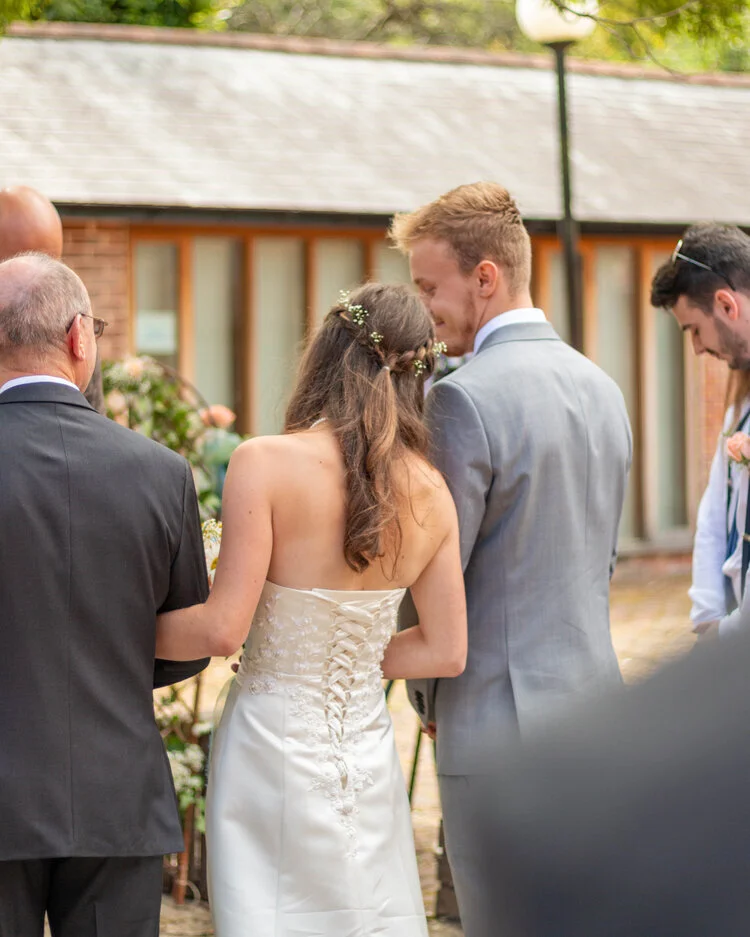 A bride and groom at their wedding ceremony, facing each other and holding hands, surrounded by friends and family outdoors.