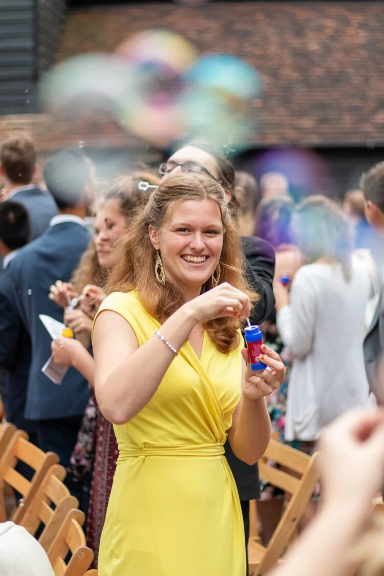 A woman in a yellow dress is blowing bubbles at an outdoor gathering with people in the background.
