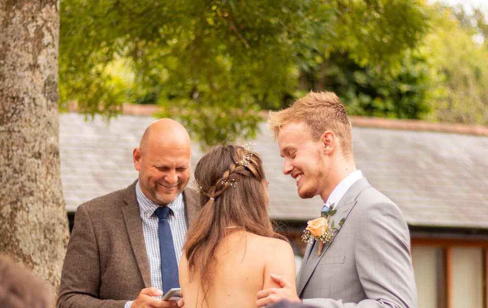 A couple in wedding attire sharing a joyful moment outdoors, with a man in a brown suit and a woman with long hair, and another man in a gray suit smiling nearby.