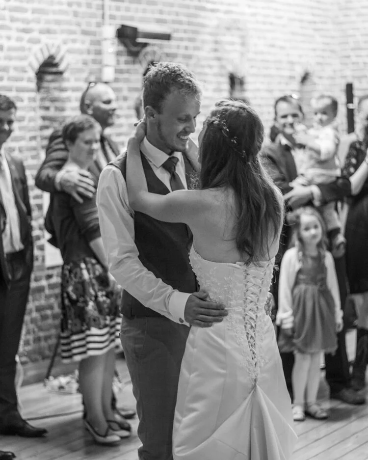 A couple dancing at a wedding, surrounded by guests in a venue with brick walls.