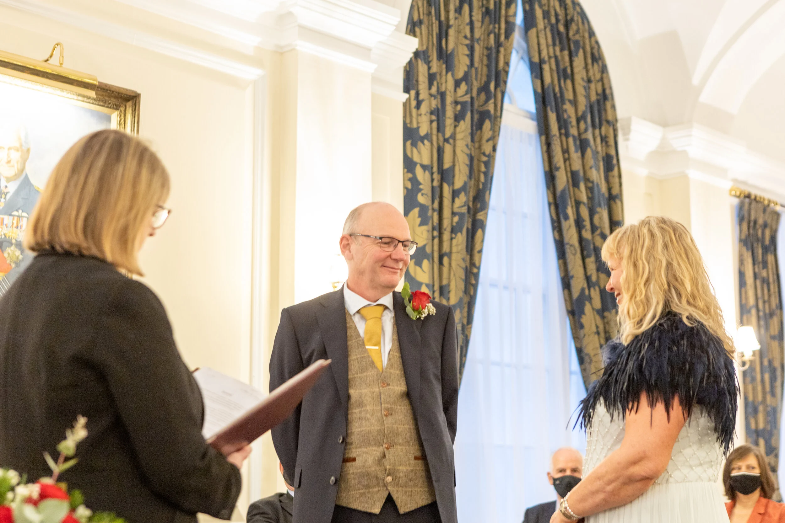 An older man in a suit and yellow tie is smiling at an older woman in a white dress with black feathered shoulders during a formal ceremony, with a woman reading from a document nearby and people seated in the background.