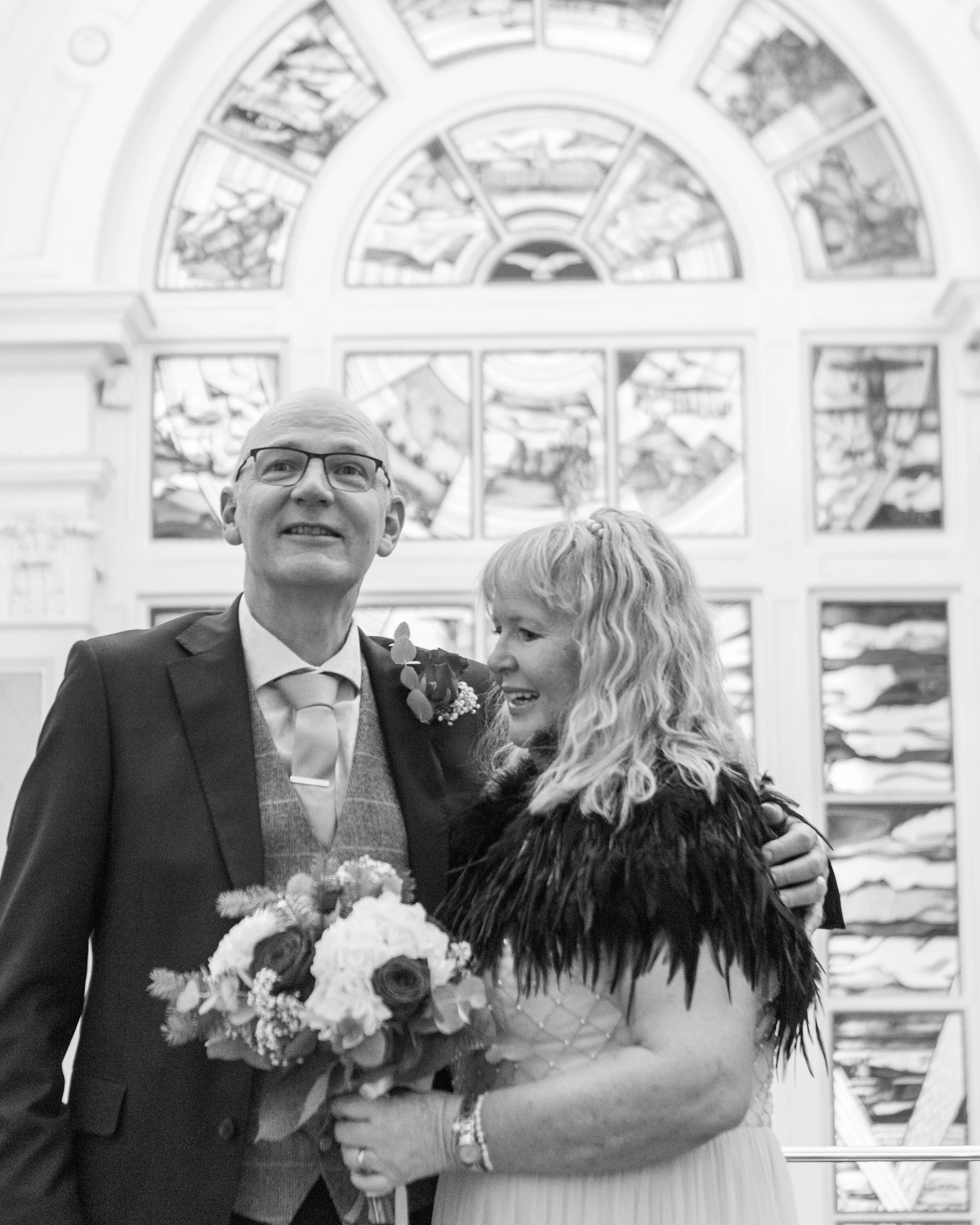 A black-and-white photo of a man and woman on their wedding day, standing in front of a stained-glass window. The man is wearing a suit and glasses, holding a bouquet, and smiling. The woman has curly hair, wearing a dress with feathered accents, and