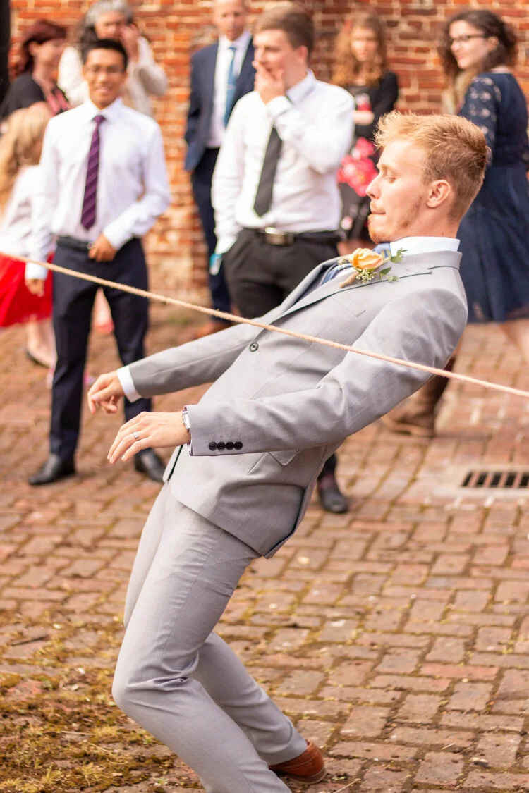 A man in a light gray suit is playing a game of tug-of-war with a rope during a wedding celebration, surrounded by guests watching and smiling.