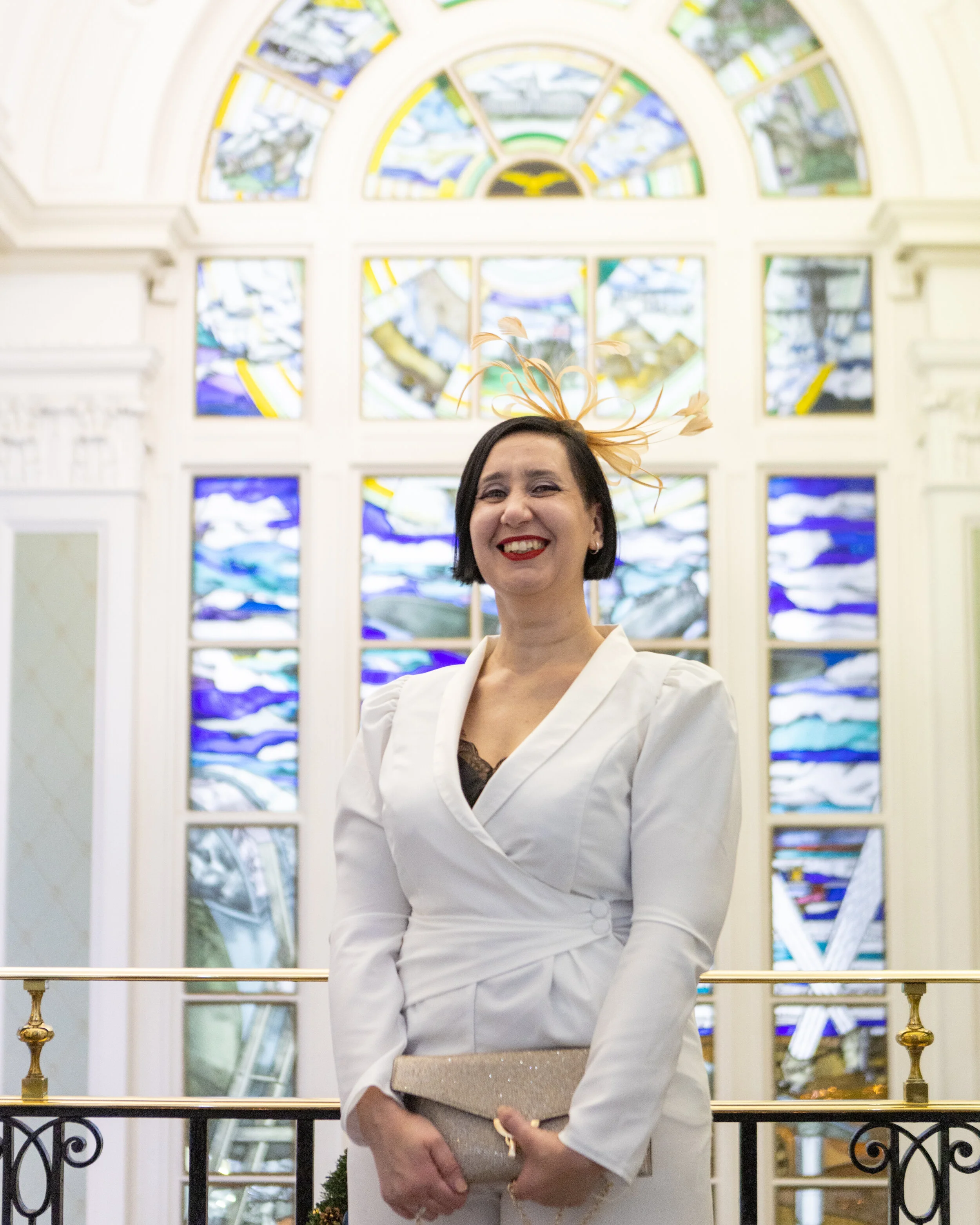 Woman in white dress with a feathered fascinator standing in front of colorful stained glass windows, holding a beige clutch, smiling.