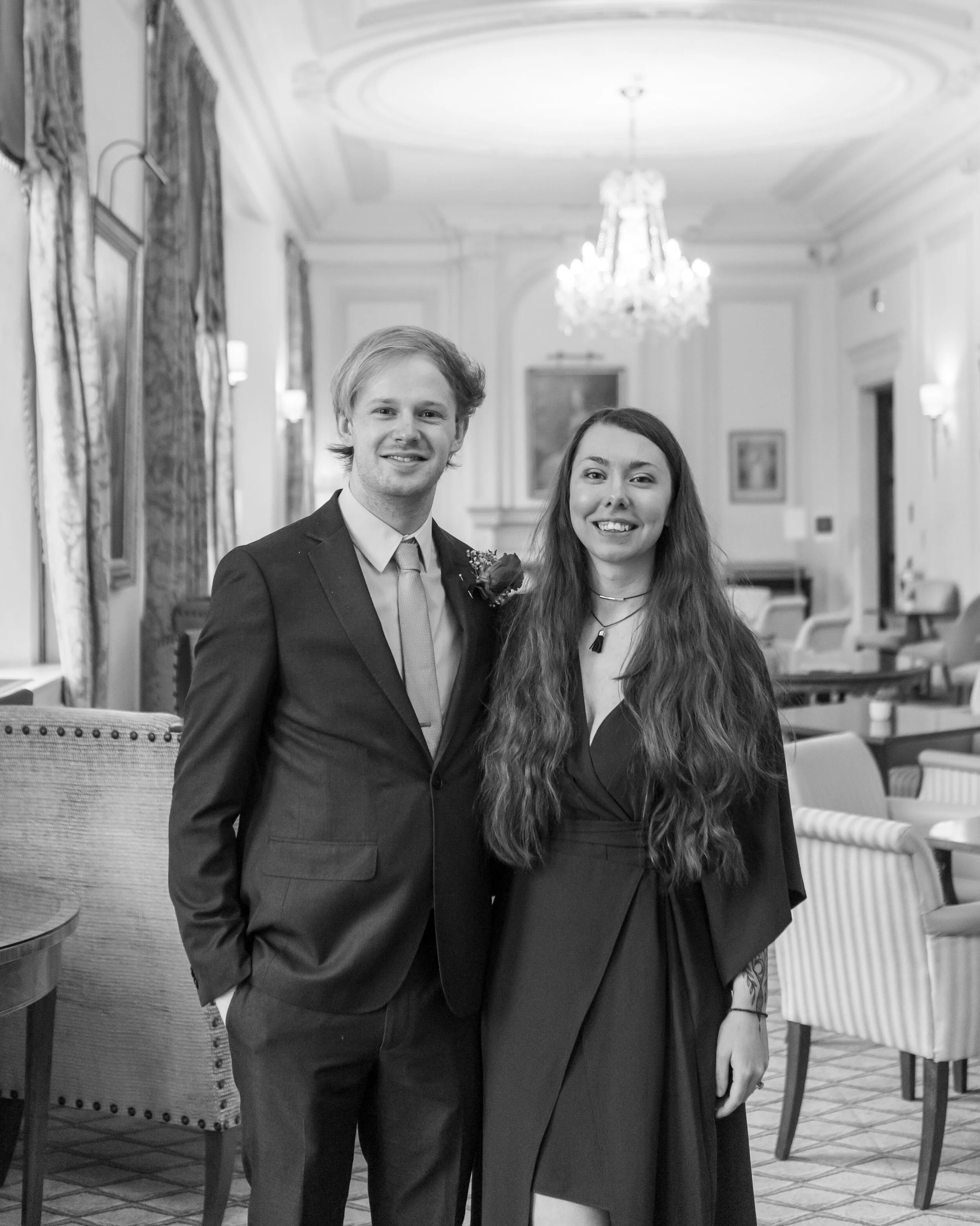 A young man and woman in formal attire standing inside a decorated room, smiling at the camera.
