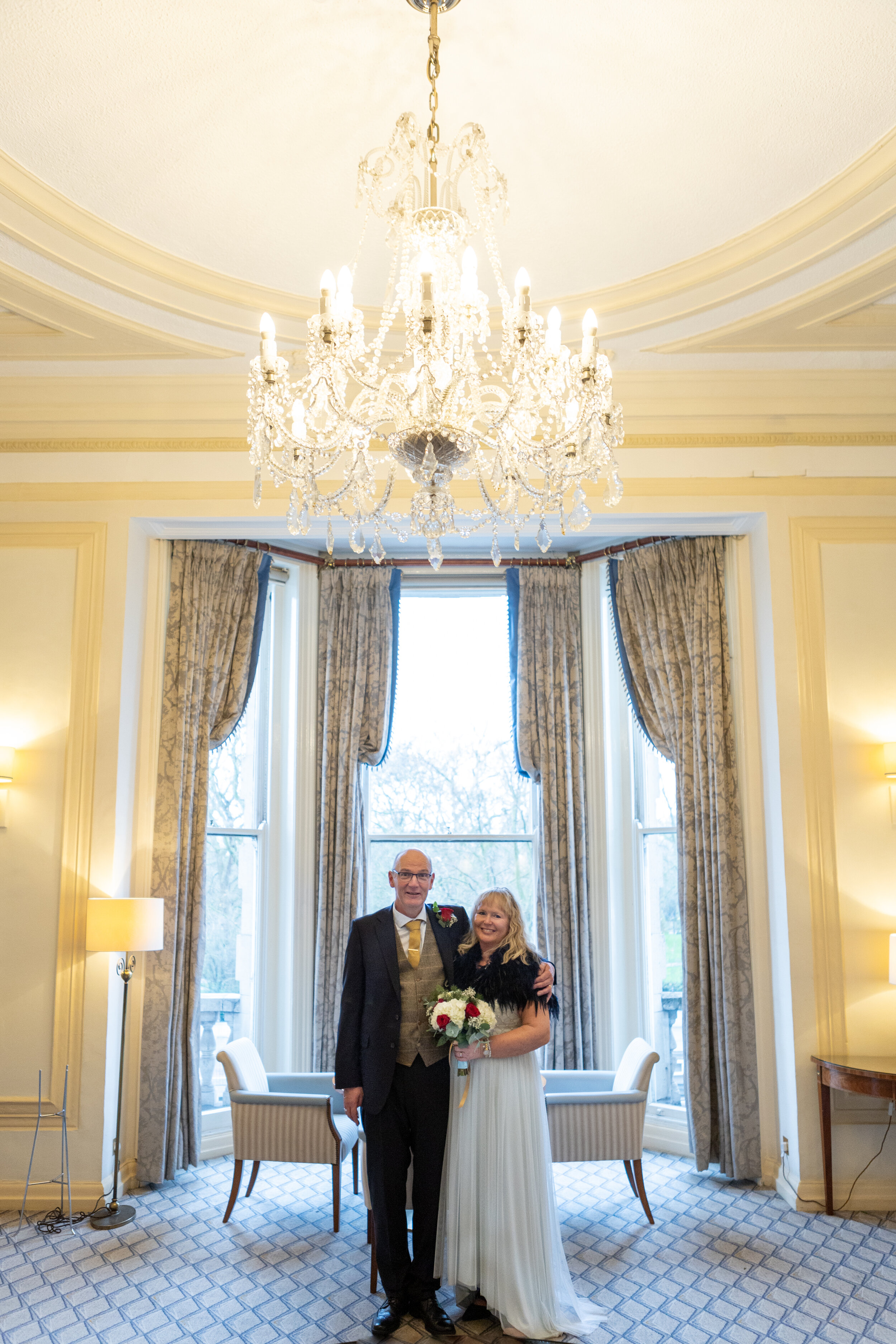 A bride and groom standing together inside a well-lit room with elegant curtains, a large chandelier, and patterned carpet.