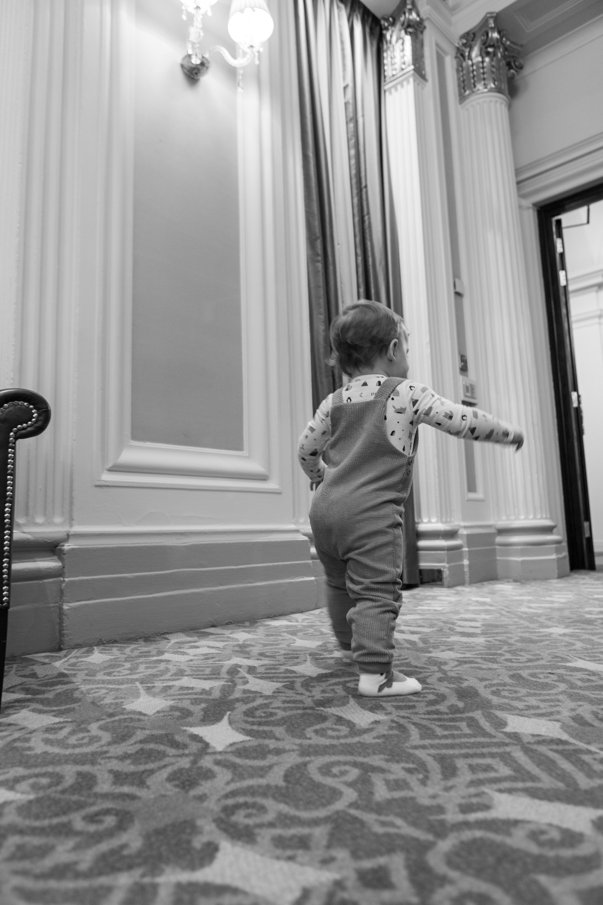 A young child walking on a patterned carpet in a luxurious, ornate room with large columns, detailed molding, curtains, and a chandelier.