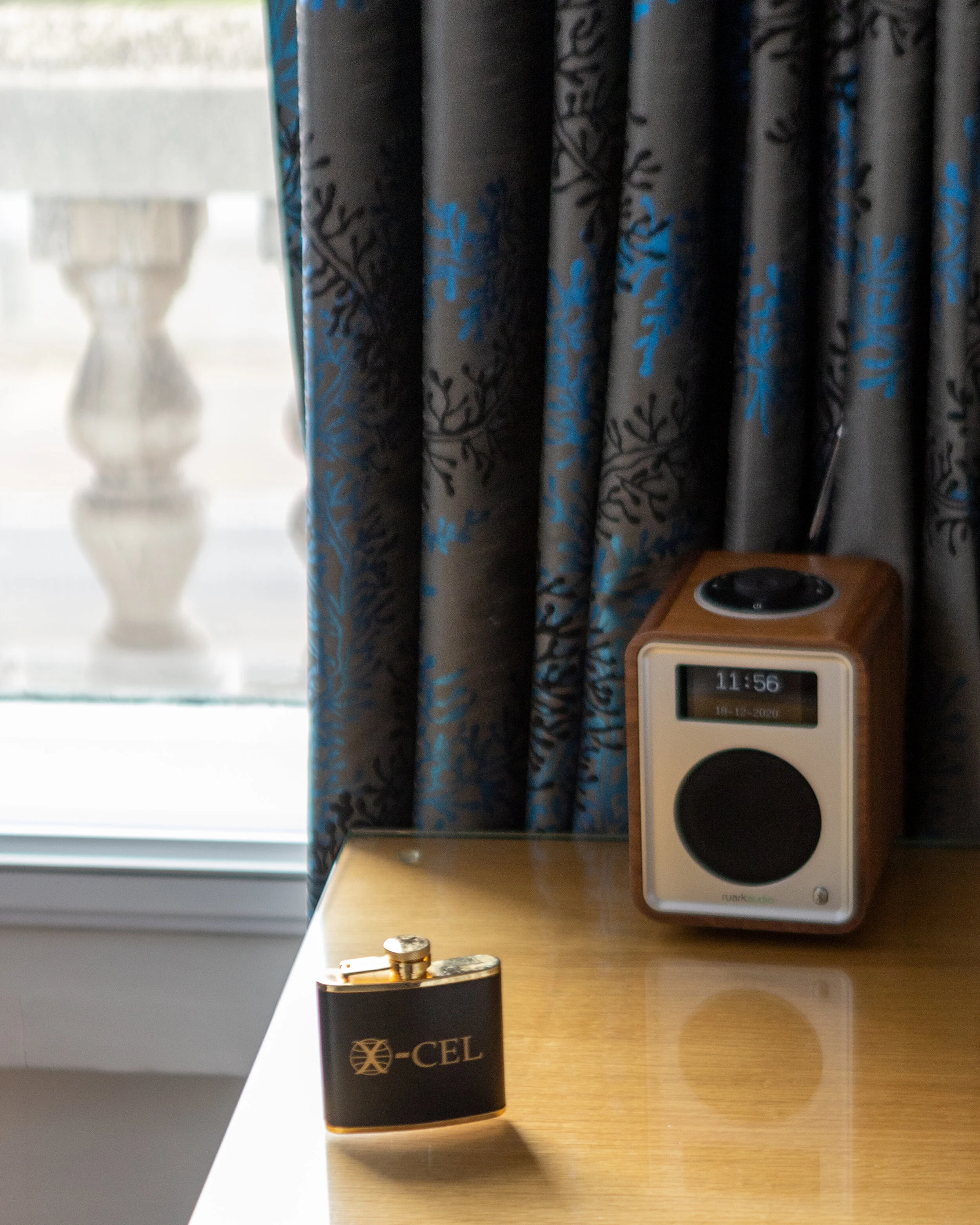 A small black X-CEL branded flask on a wooden surface, a digital clock showing 11:56 in a wooden speaker, and a window with blue and black patterned curtains.