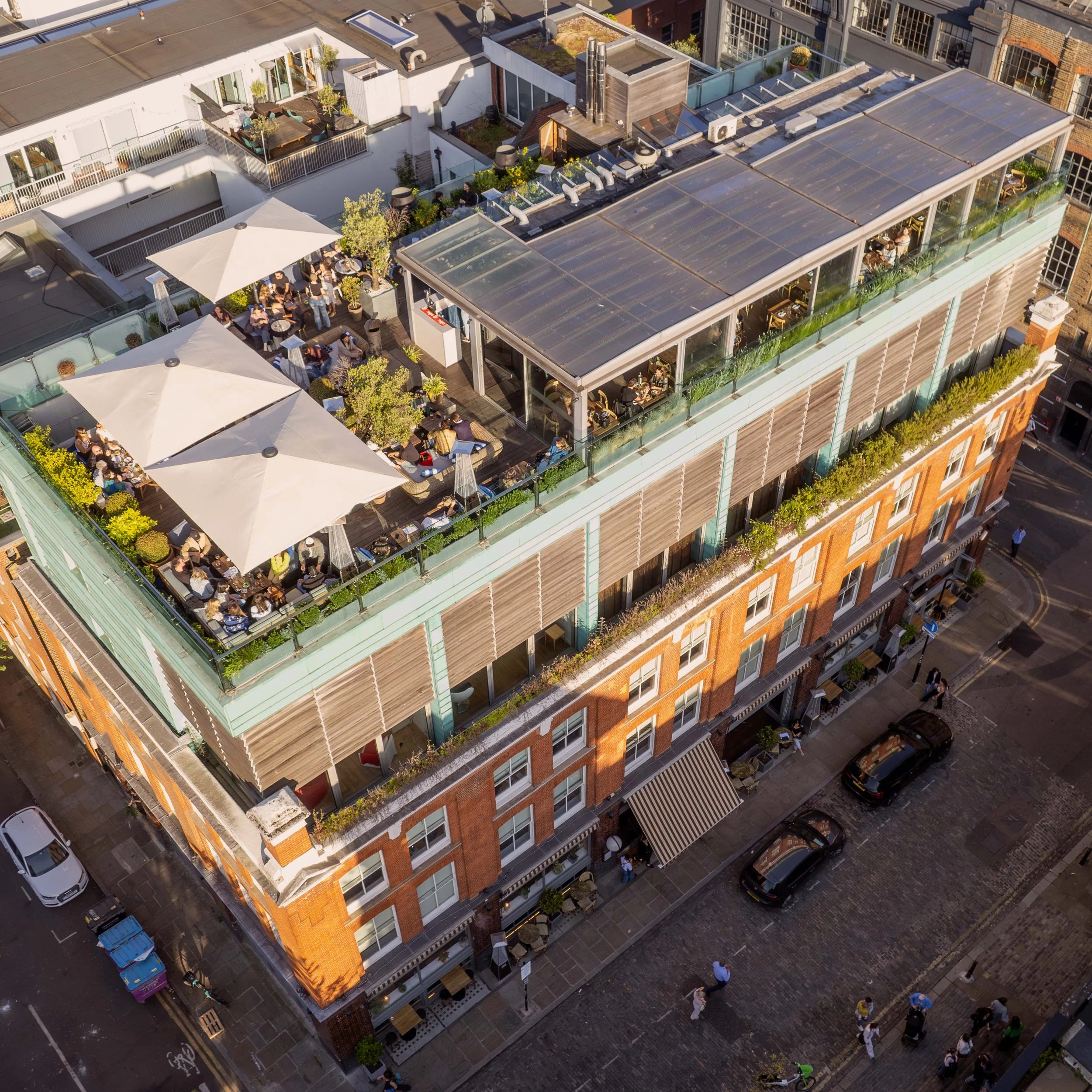 Aerial drone view of a multi-story building in Shoredit with a rooftop restaurant and outdoor seating, with many people dining under umbrellas and on a partially enclosed terrace, in an urban area with parked cars and pedestrians on the street below.