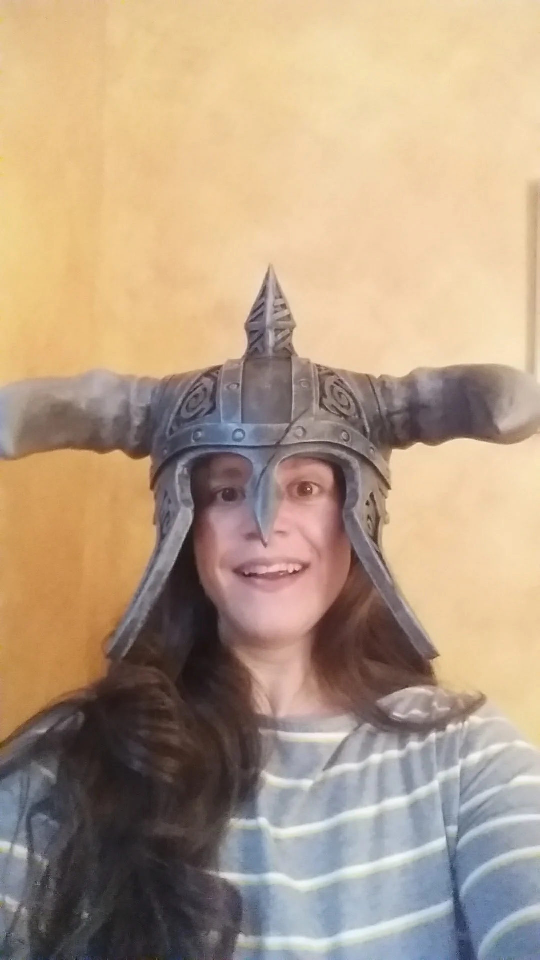 Woman with long brown hair smiling at camera, wearing a Viking-style helmet with large horns and intricate designs, set against a plain yellow background.