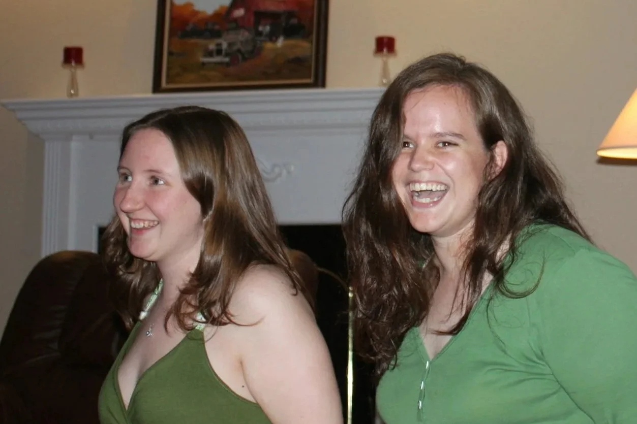 Two women with long brown hair, smiling and laughing, standing in a living room with a fireplace and framed pictures on the wall, both wearing green tops.