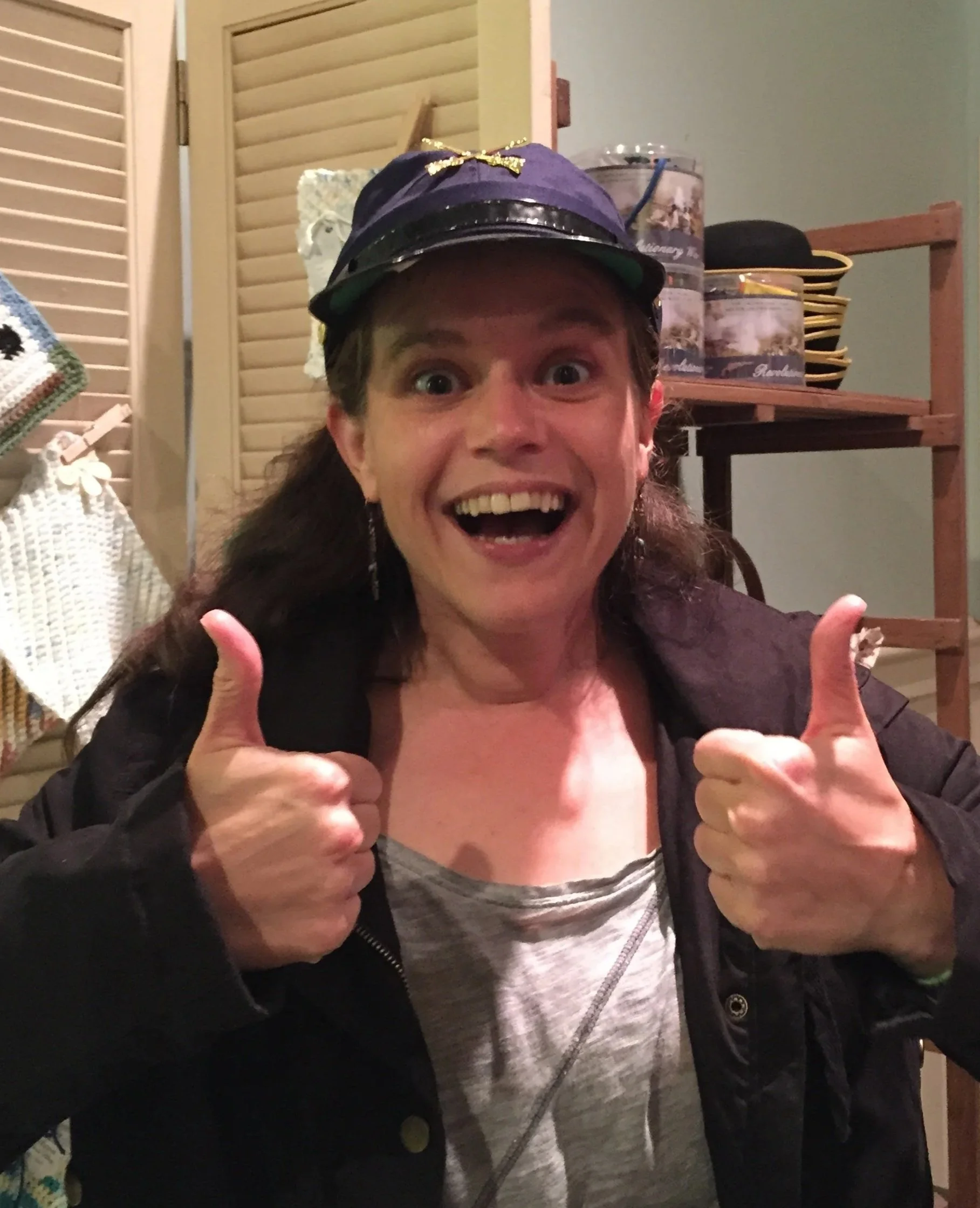 A woman smiling with her mouth open and giving a thumbs-up. She is wearing a black jacket, a gray shirt, and a purple helmet with a golden bow on top. The background includes a wooden shelf with bowls, a lidded box, and some black hats, with a wooden shutter and lace decoration on the wall.