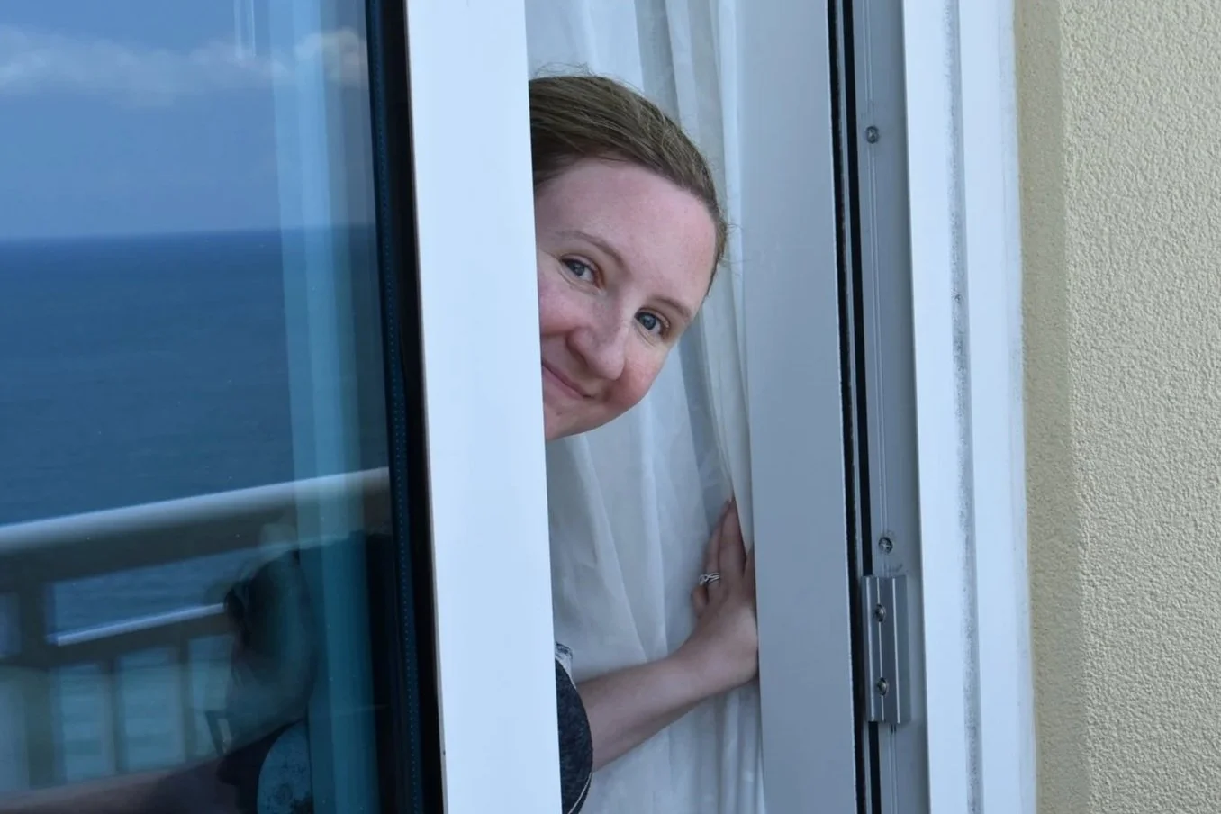 A woman peeking out of a window, smiling, with her hand resting on the window frame.