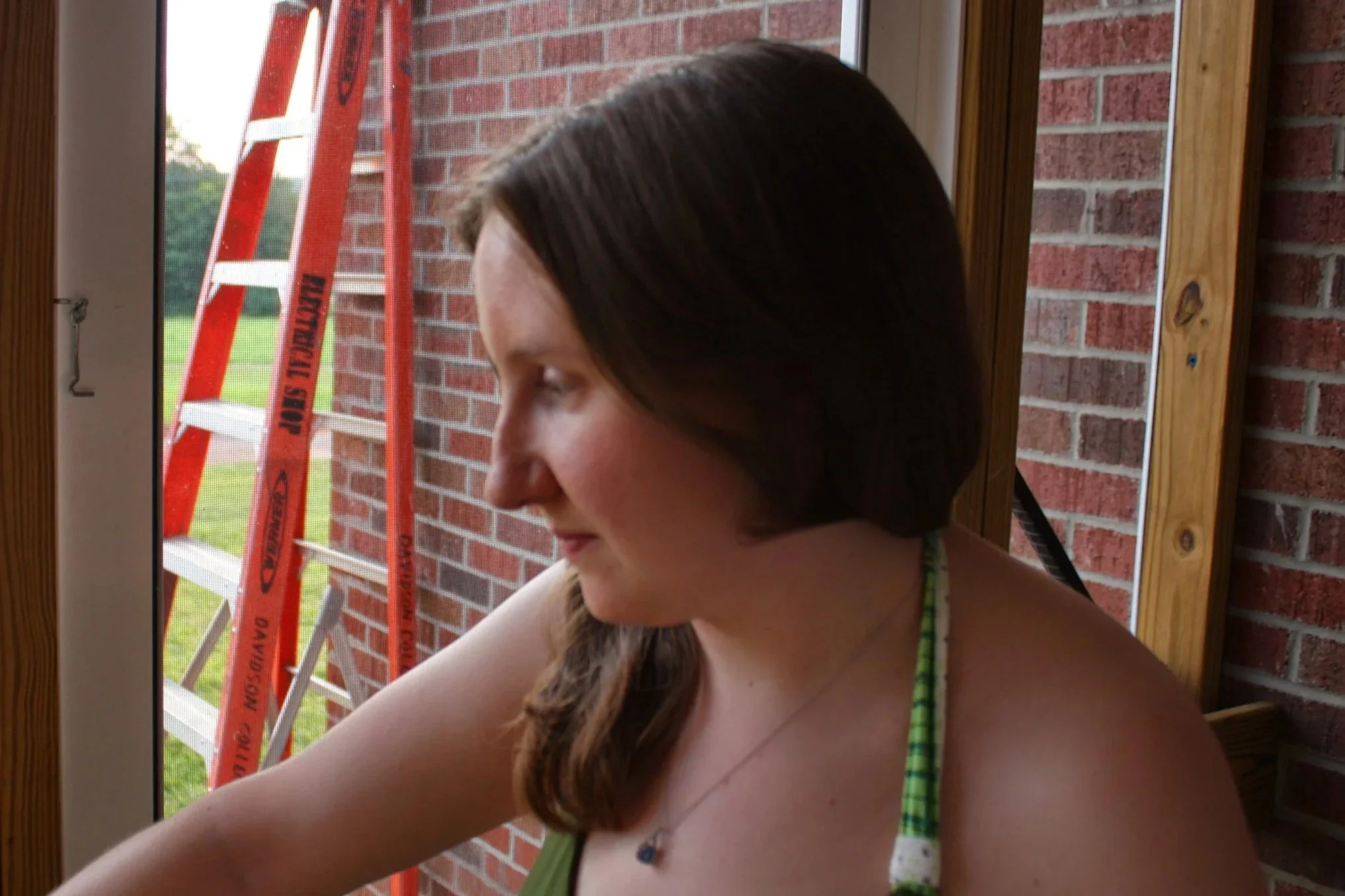 Profile of a young woman with brown hair and a green striped tank top, standing by a window with a brick wall and an orange ladder outside.