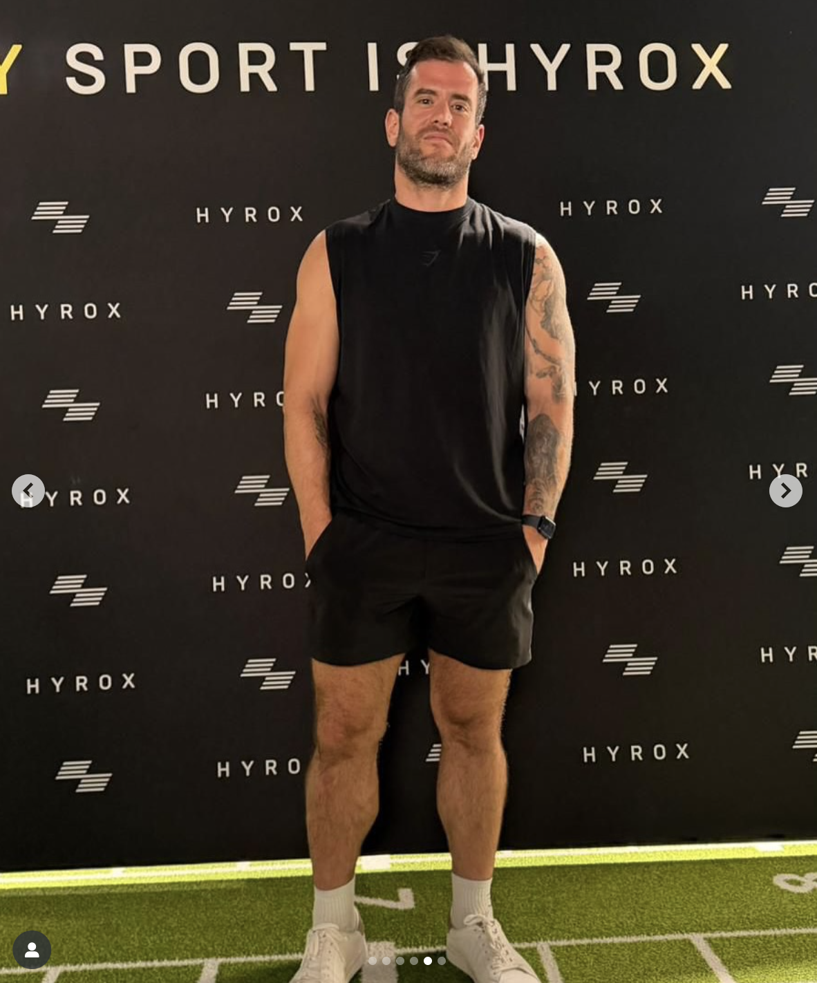 Man standing in front of a black backdrop with 'HYROX' and logo, wearing black athletic sleeveless shirt, shorts, white sneakers, and a smartwatch, with tattoos on arms.