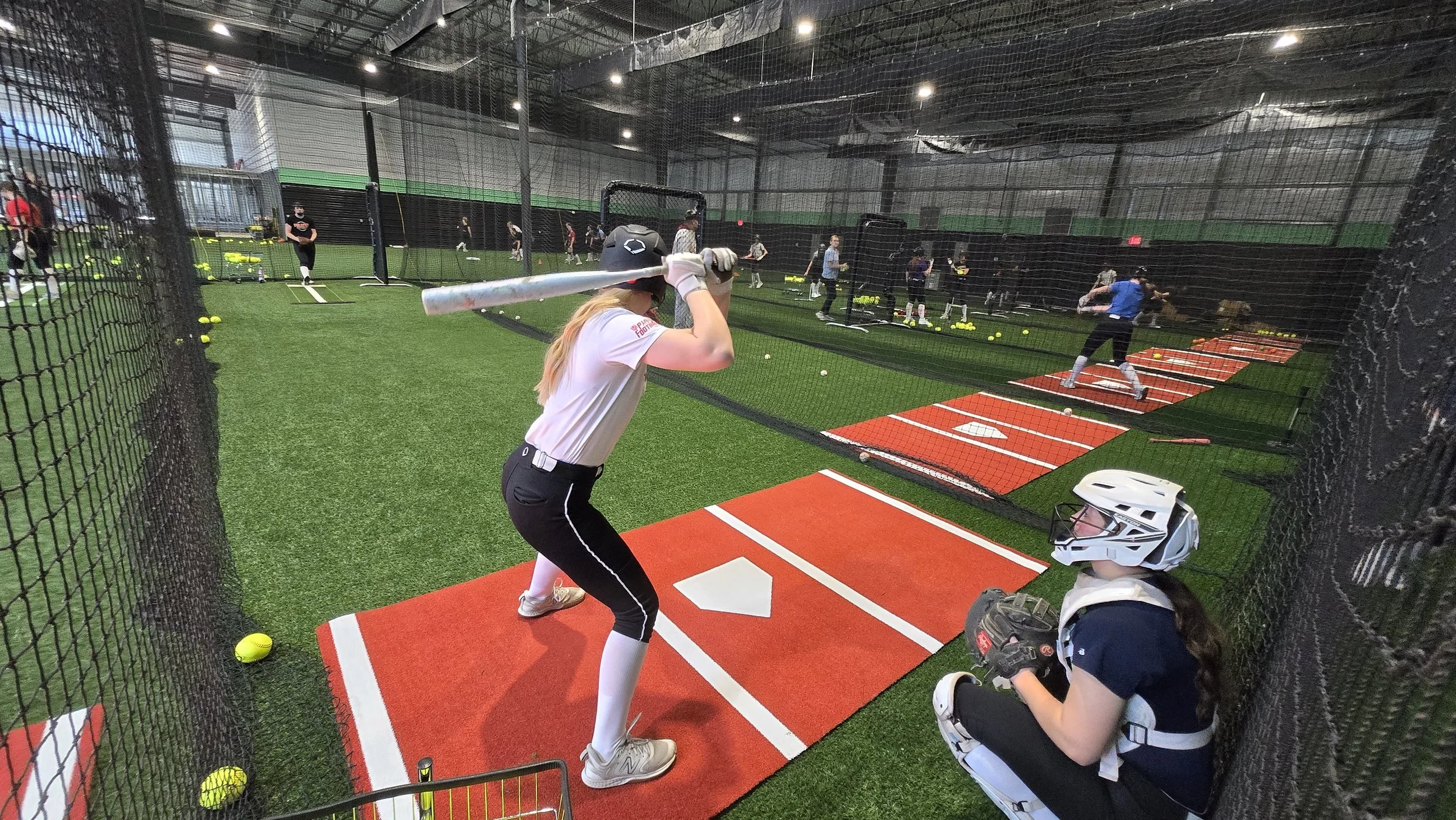 PWR FASTPITCH An indoor baseball training facility with multiple players practicing batting, a catcher in protective gear kneeling behind her, and several others practicing in the background.