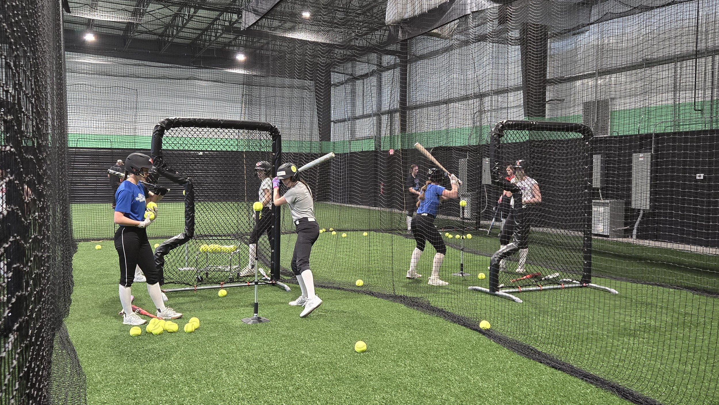 PWR FASTPITCH Women practicing baseball in an indoor training facility with protective netting, turf flooring, and scattered tennis balls, wearing helmets and athletic clothing.