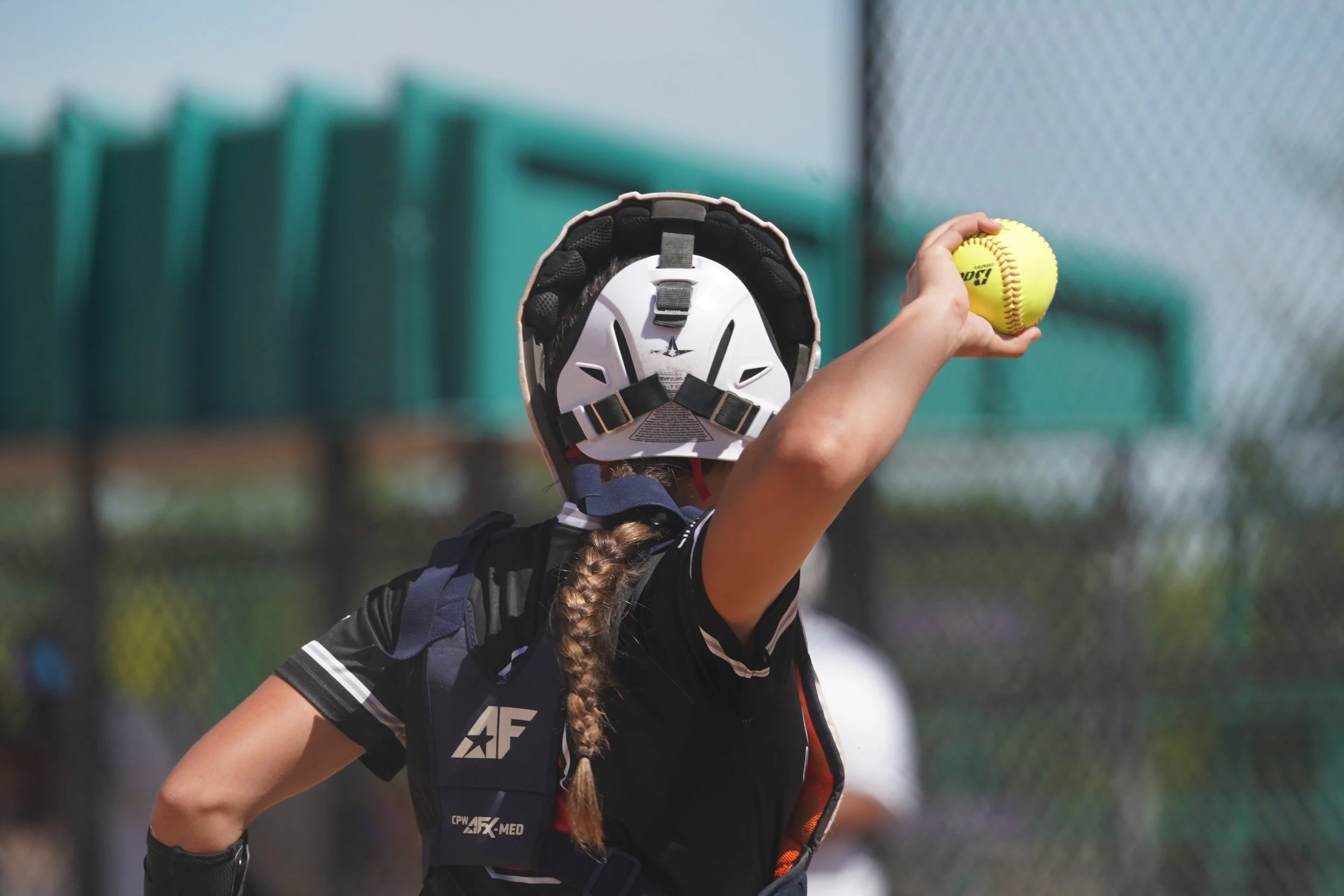 A softball player wearing a helmet, black jersey, and a protective vest is preparing to throw a yellow softball on a field with a chain-link fence and green bleachers in the background.