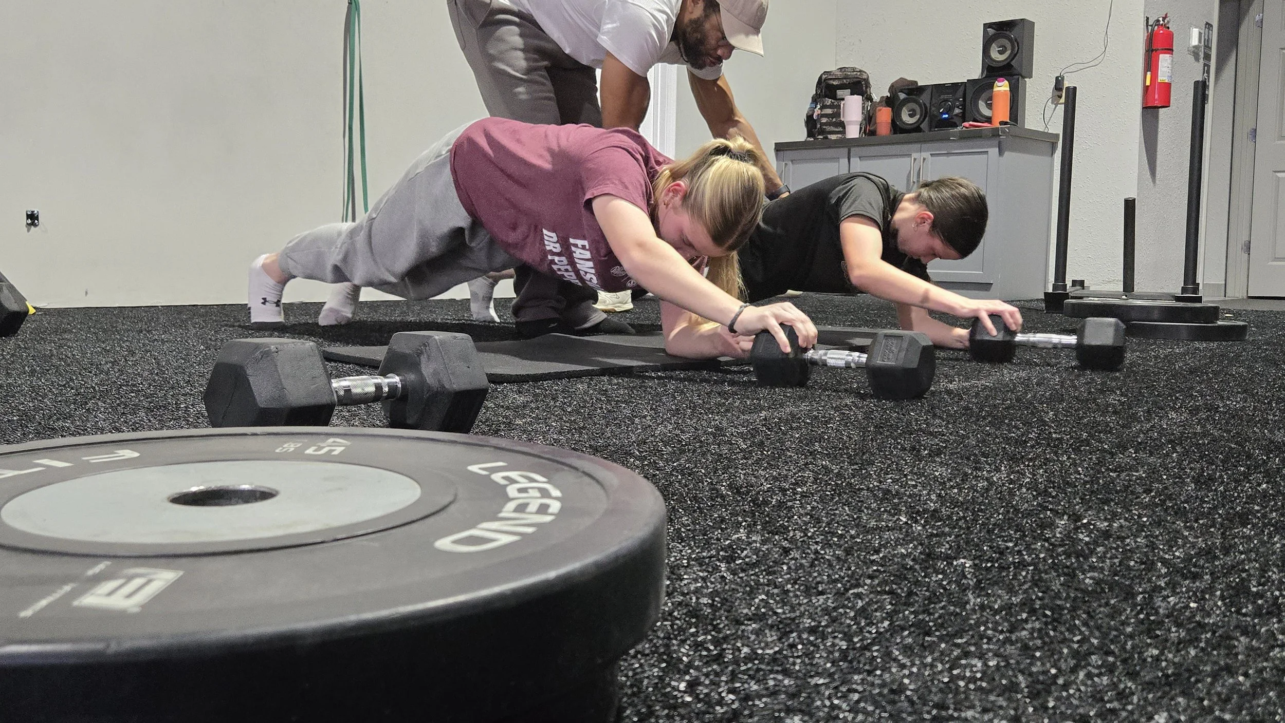 PWR FASTPITCH Women performing planks with dumbbells during a workout session on a gym floor.