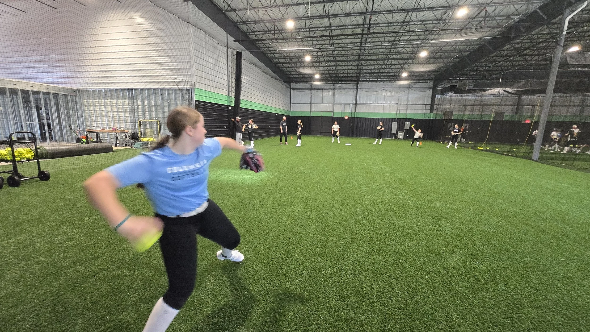 PWR FASTPITCH Indoor sports training session with young female athlete in foreground throwing a softball, others practicing in the background on artificial turf field