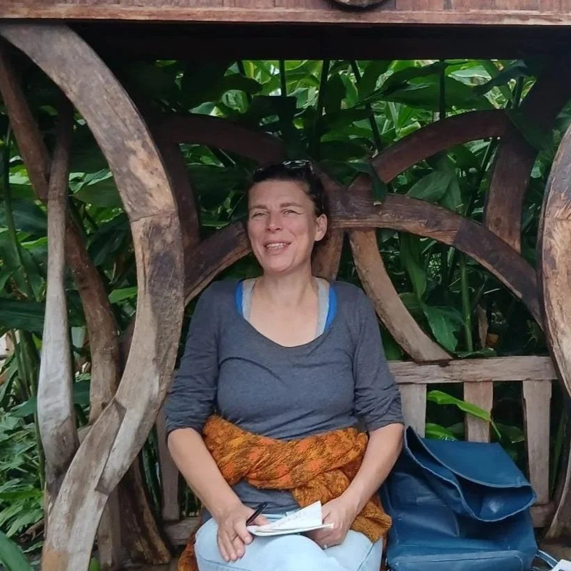 Vrouw in een bruin pak die binnen staat, glimlacht, een koffiebeker en een tas vasthoudt, met planten op de achtergrond.