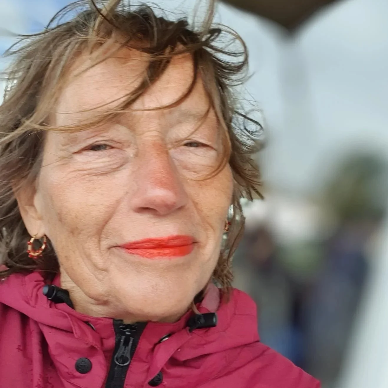 Close-up van een oudere vrouw met warrig bruin haar en rode lipstick, draag een rode jas.