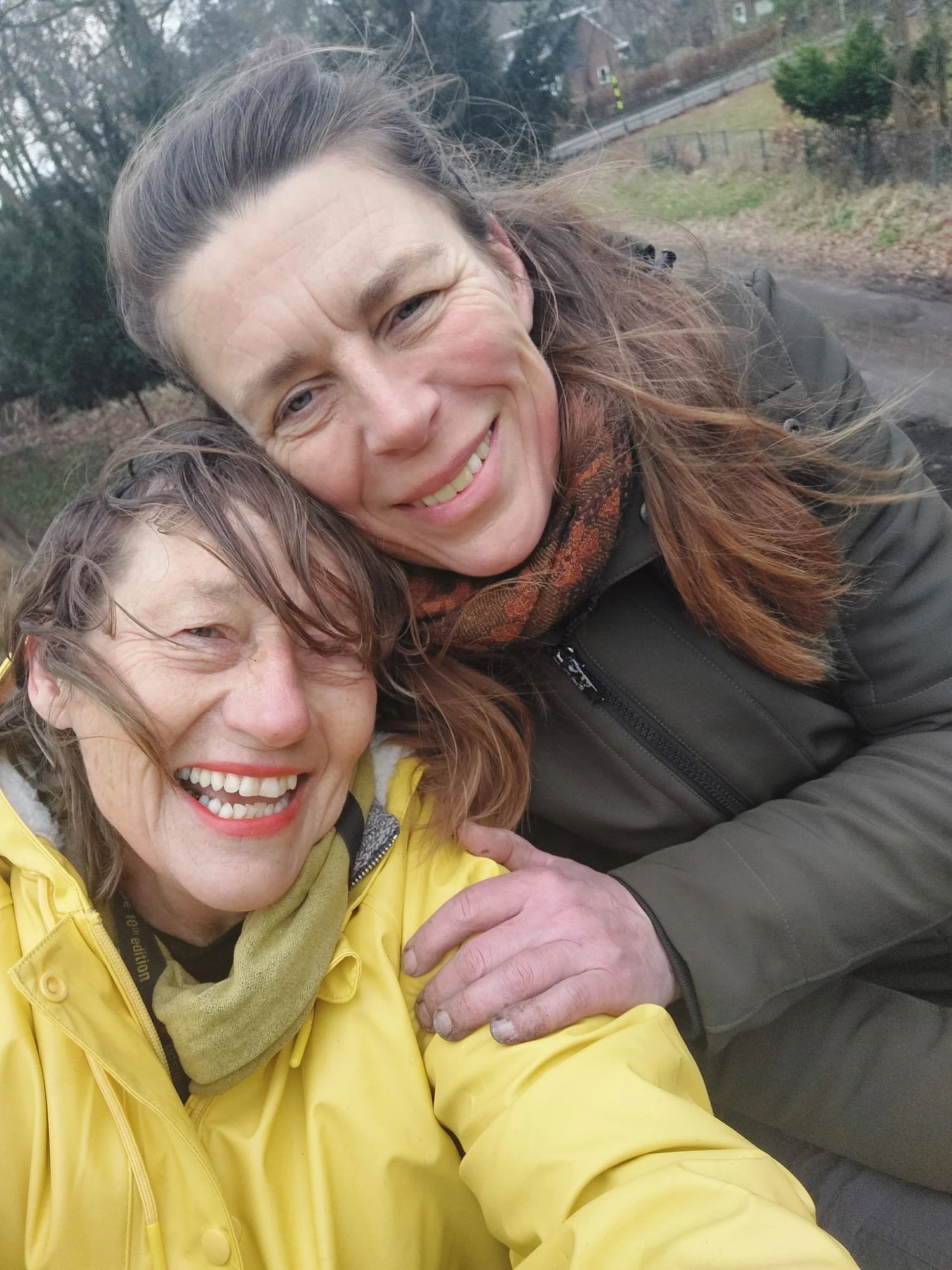 Twee lachende vrouwen die een selfie nemen buiten, één in een gele jas en de ander in een zwarte jas, met een weg en bomen op de achtergrond.