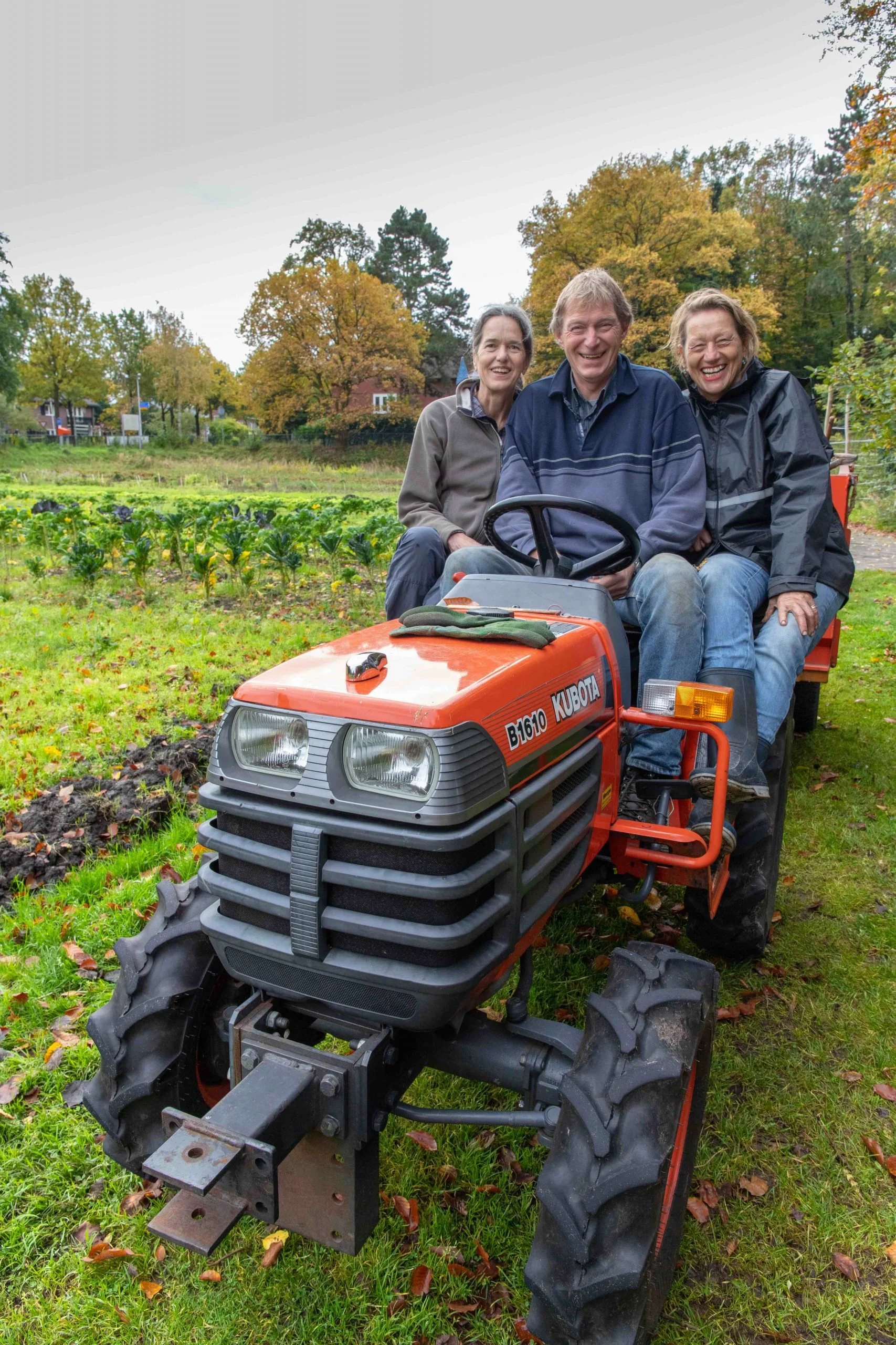 Drie mensen zitten op een kleine tractor in een tuin met gewassen, herfstkleuren en bomen op de achtergrond.