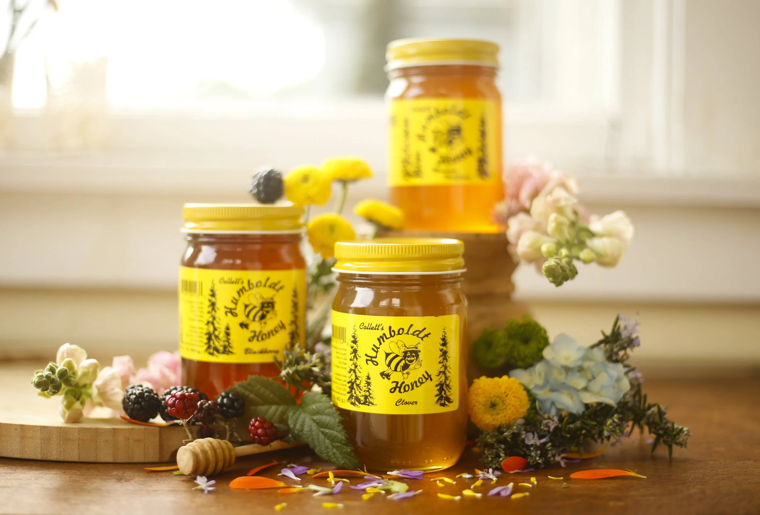 Three jars of honey labeled 'Humboldt Honey' with yellow labels featuring a bear and trees, surrounded by flowers and berries on a wooden surface near a window.