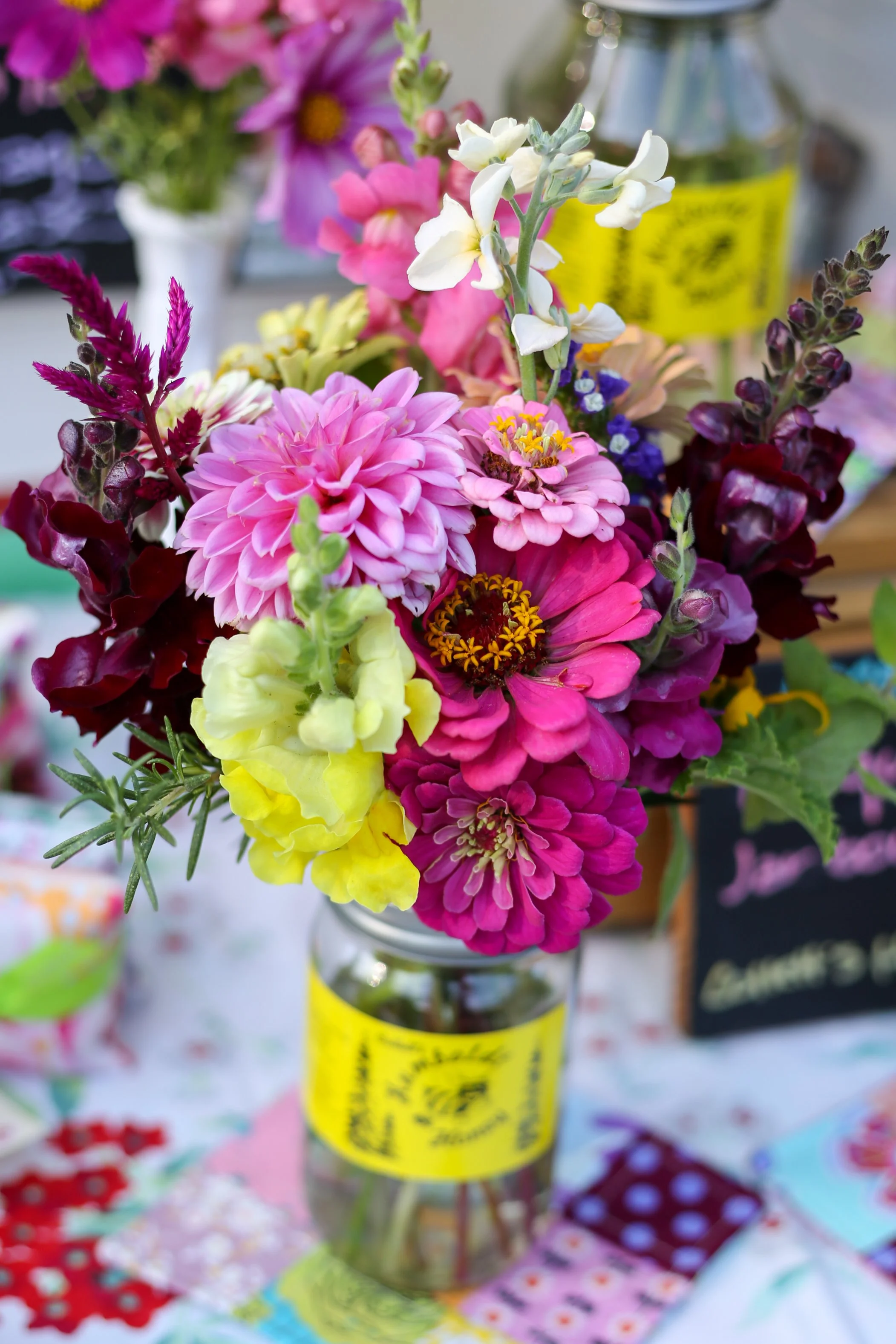 Colorful bouquet of mixed flowers in a glass jar on a table, with a yellow label on the jar and various patterned cloths underneath.