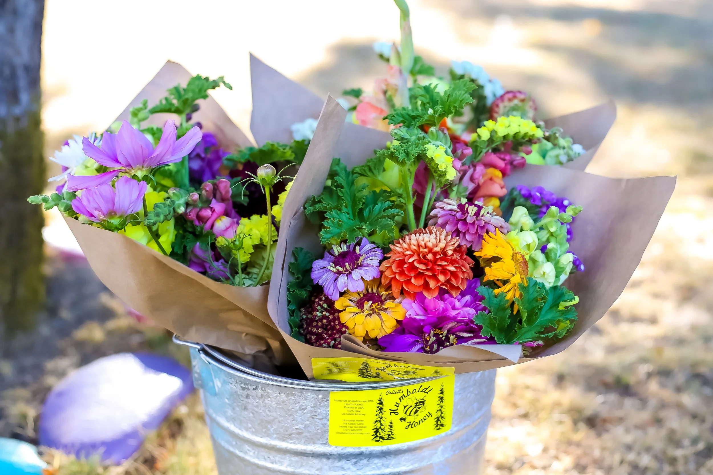 Colorful bouquet of various flowers wrapped in brown paper, placed in a silver bucket with a yellow label, outdoors on a sunny day.