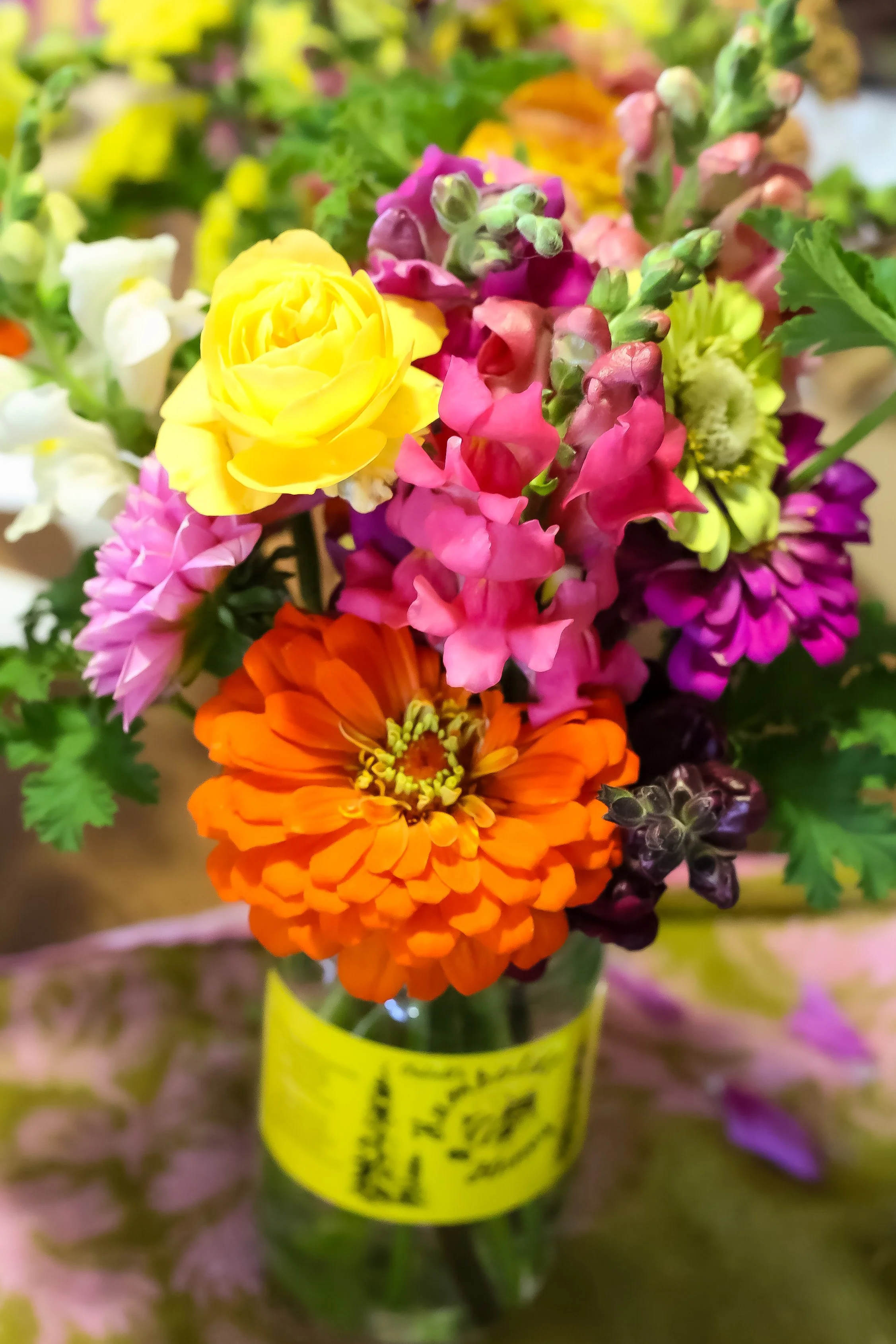 Colorful bouquet of various flowers including yellow, pink, orange, purple, and white blossoms in a glass jar with a yellow label.