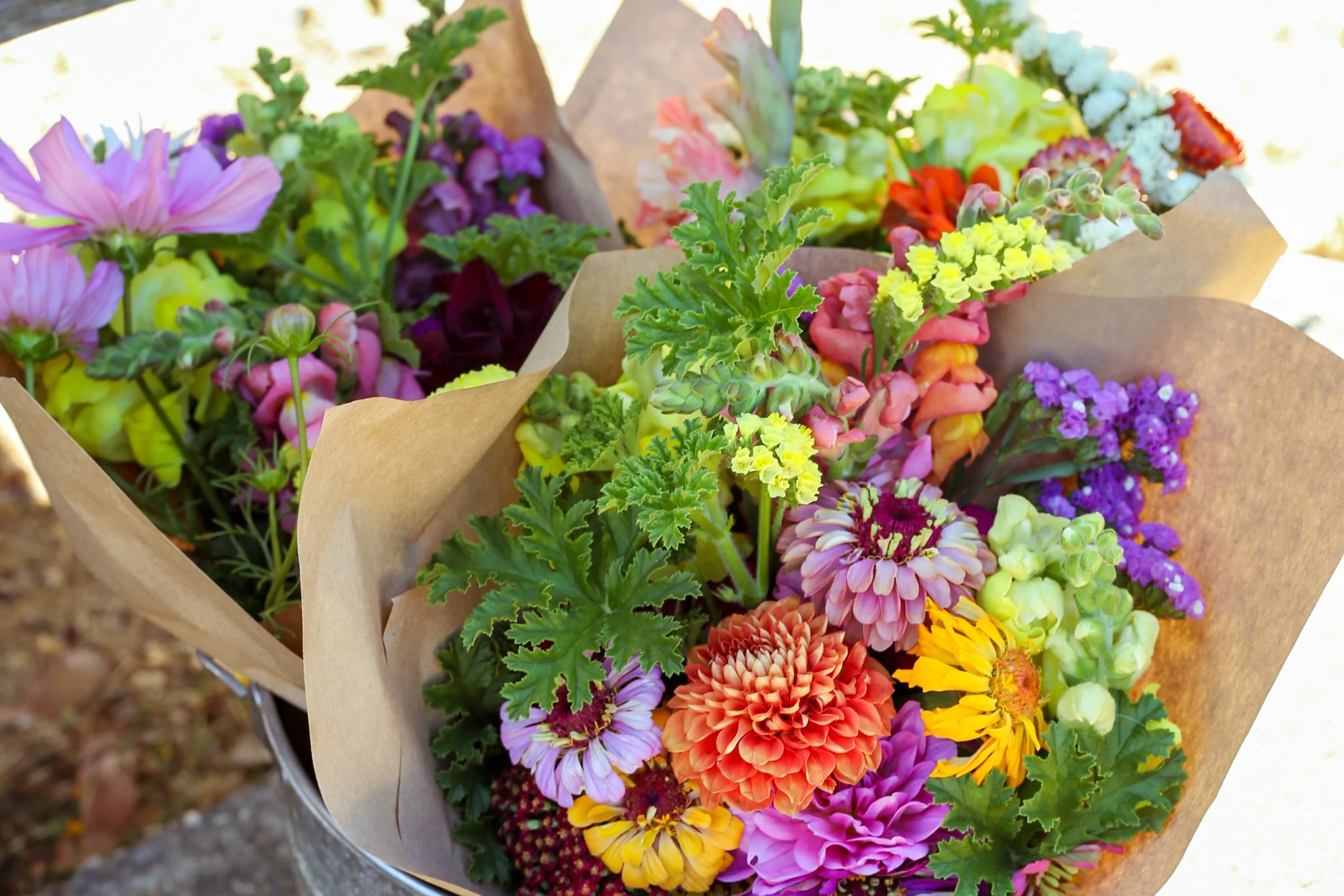 Colorful bouquets of fresh flowers wrapped in brown paper, including sunflowers, dahlias, zinnias, and other bright blossoms.
