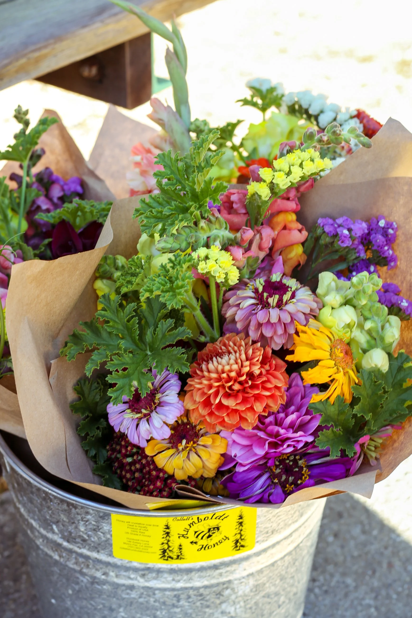 Colorful bouquet of mixed flowers including zinnias, snapdragons, and others wrapped in brown paper with a yellow label, placed in a metal bucket outdoors.