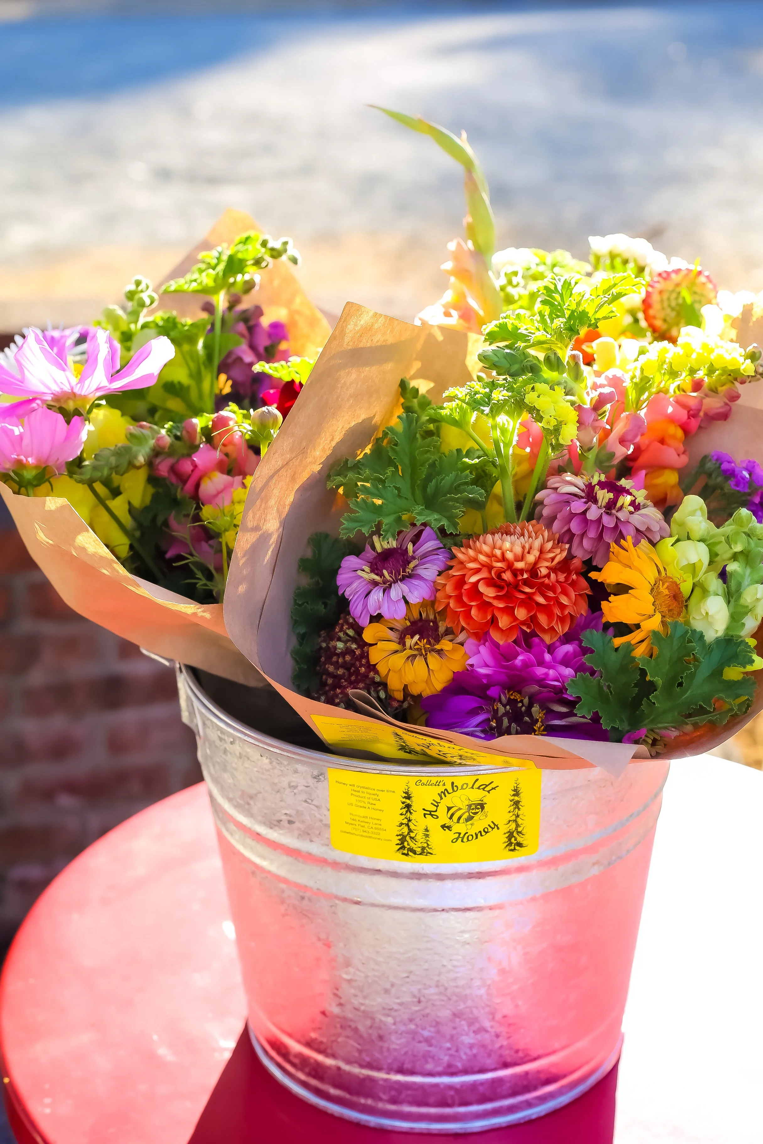 Colorful bouquet of flowers wrapped in brown paper inside a metal bucket with a yellow label. The background features a sandy beach and ocean waves.