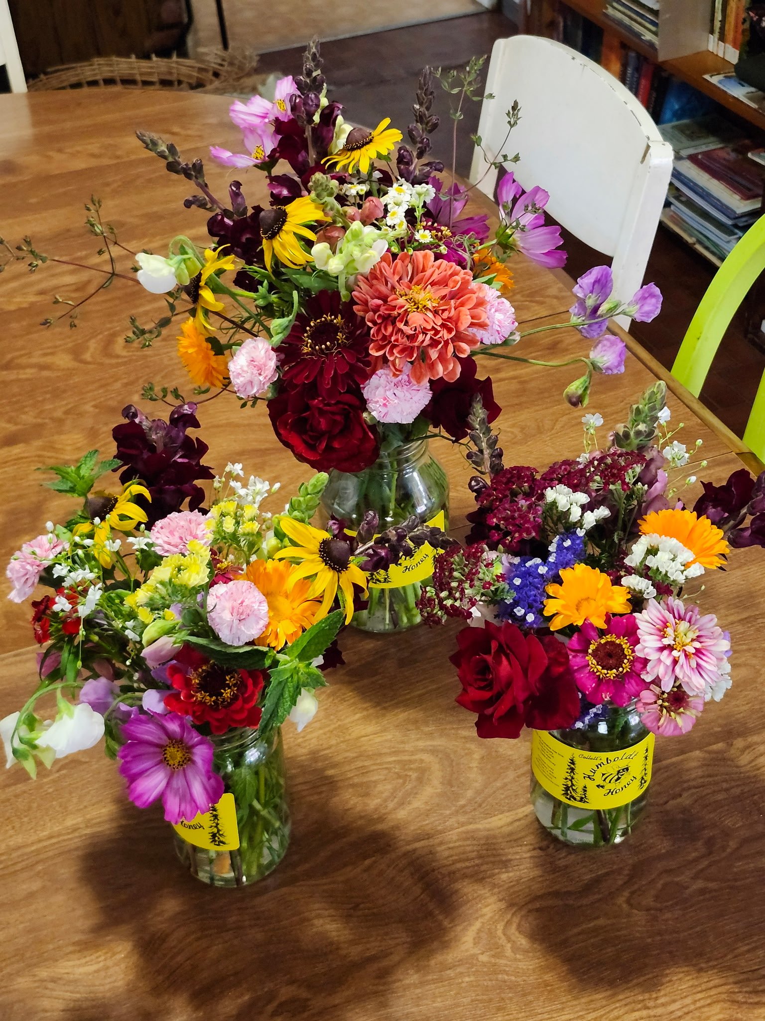 Glass jars filled with colorful garden flowers on a wooden table, with a white chair and bookshelves in the background.