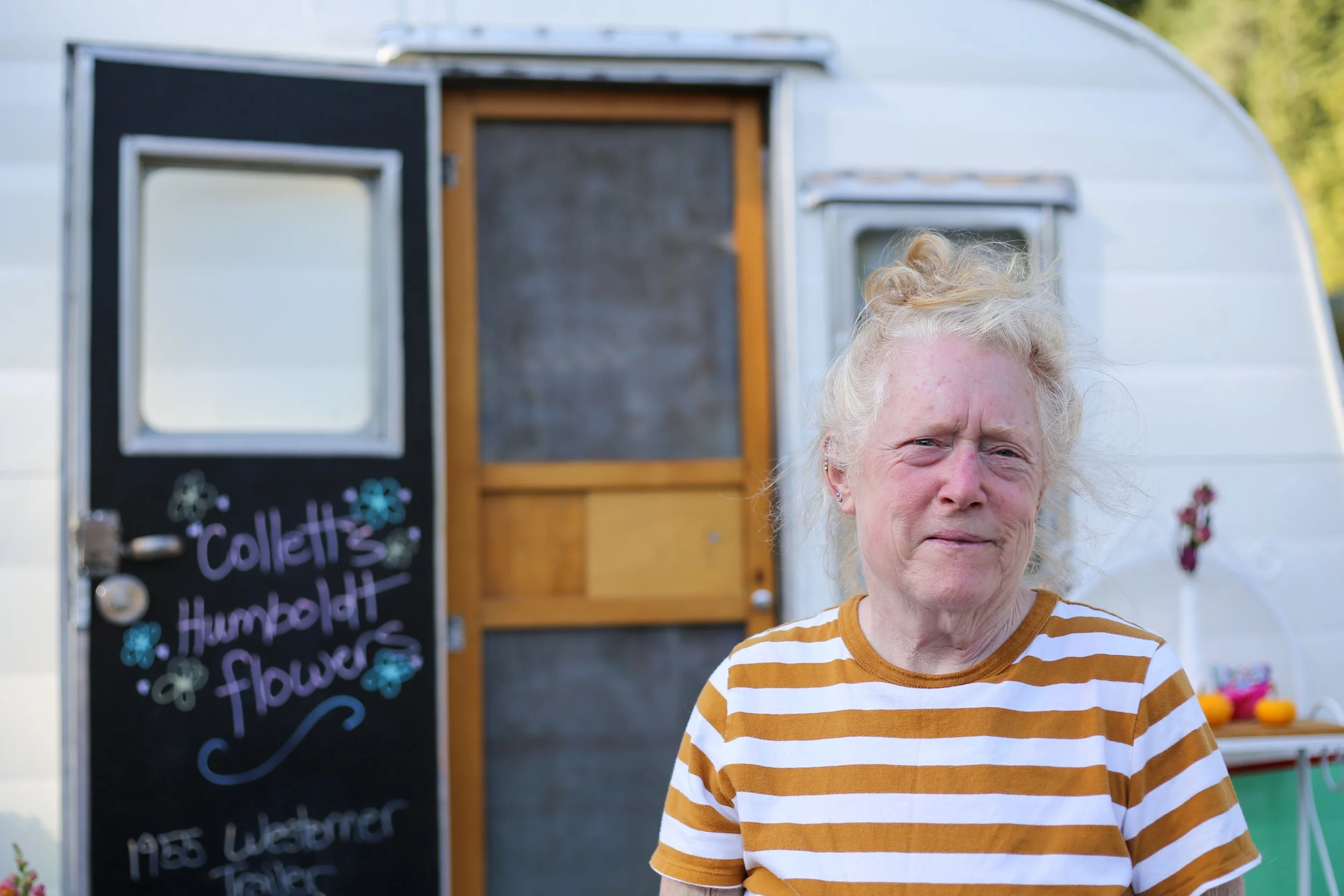 An elderly woman with blonde, curly hair wearing a yellow and white striped shirt standing outside a trailer with a black sign that reads 'Collett's Humboldt flowers' in front of a flower stand.