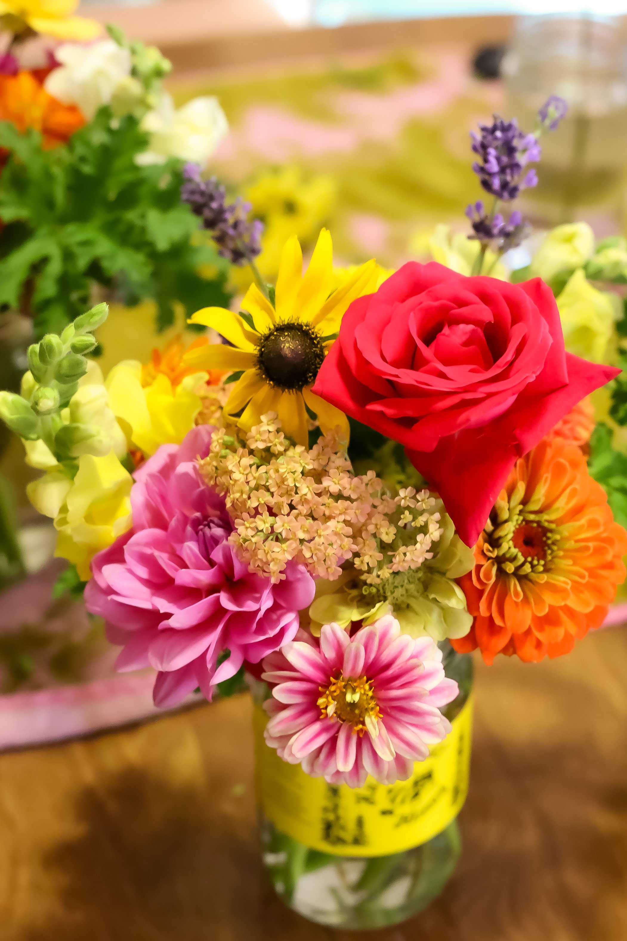 Colorful bouquet of various flowers in a glass jar, including a red rose, yellow sunflower, pink and purple dahlias, orange gerbera daisy, and lavender.