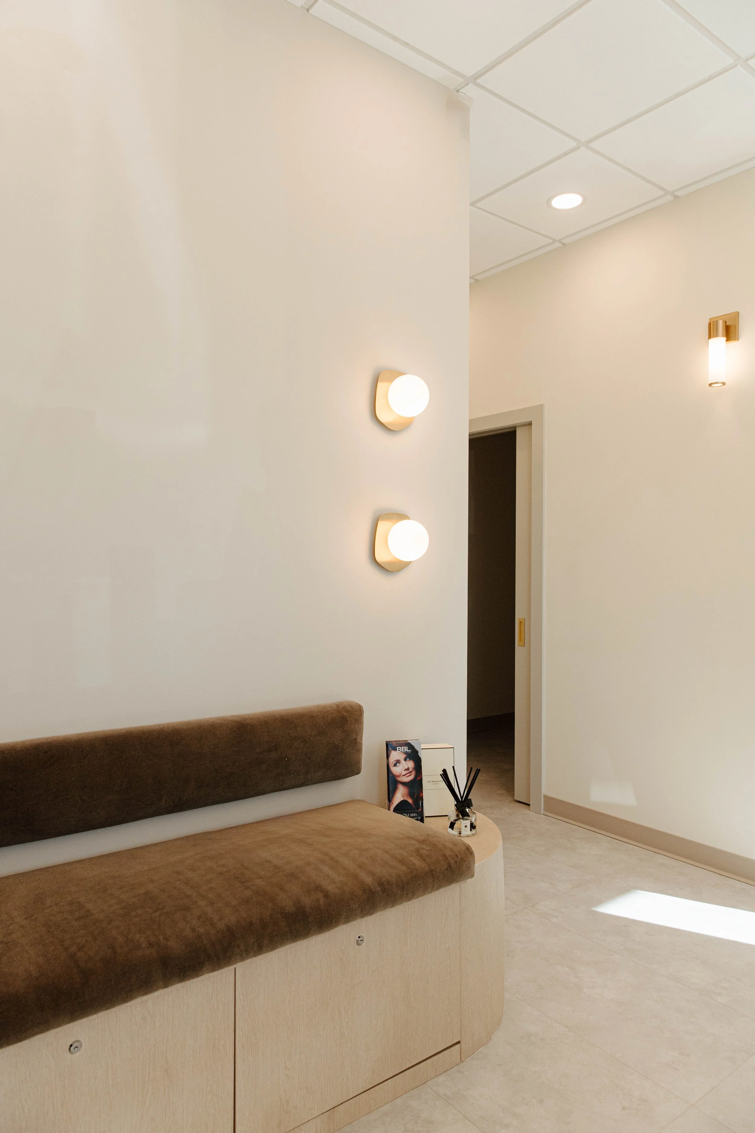 A modern waiting area with a beige bench, decorative lighting, a magazine stand, and reed diffuser on a side table, in a well-lit space with white walls and ceiling.