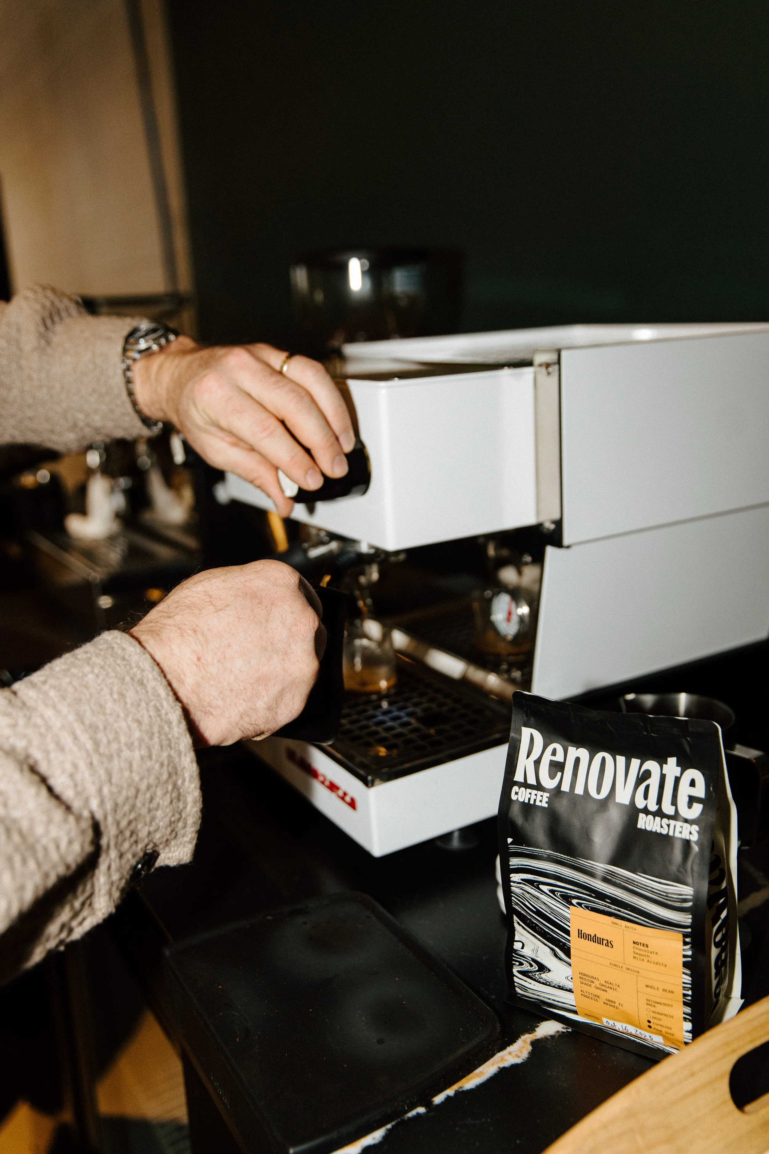 Man operating a coffee espresso machine, with a bag of Renovate Coffee Roasters Honduras coffee beans on the table nearby.