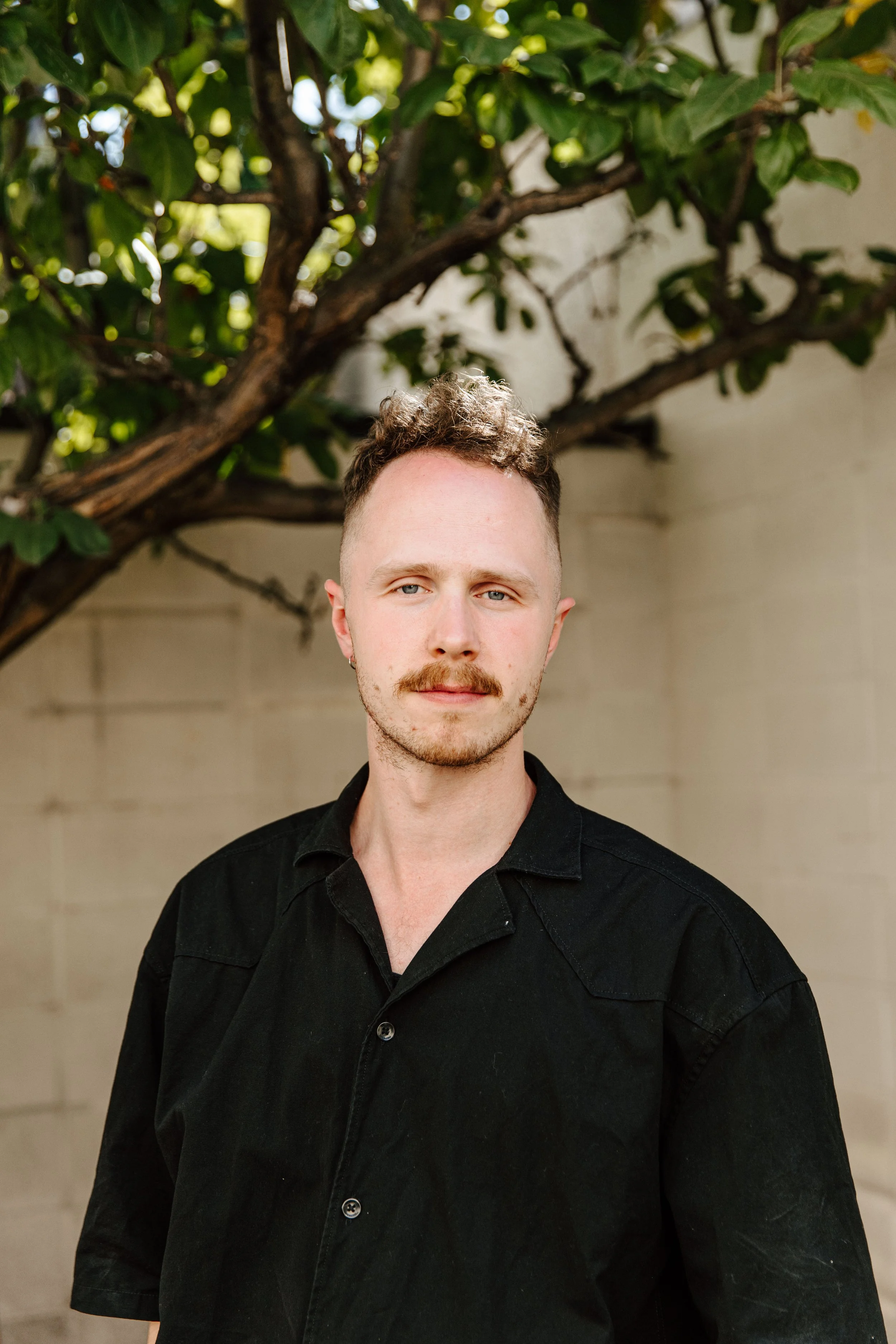 A man with short curly hair and a mustache, wearing a black button-up shirt, standing outdoors in front of a tree and beige wall.